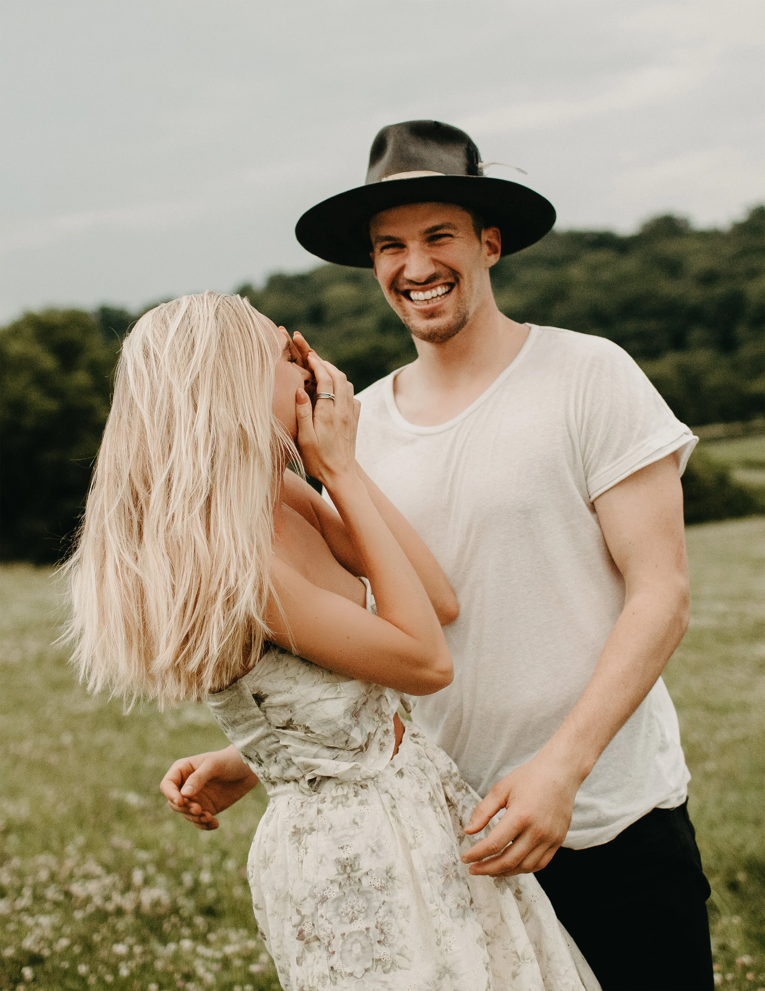 A man wearing a black wide-brimmed hat and white t-shirt is smiling, standing outdoors with a woman with long blonde hair and a floral dress, who is touching the man's face. The background features green trees and an overcast sky.