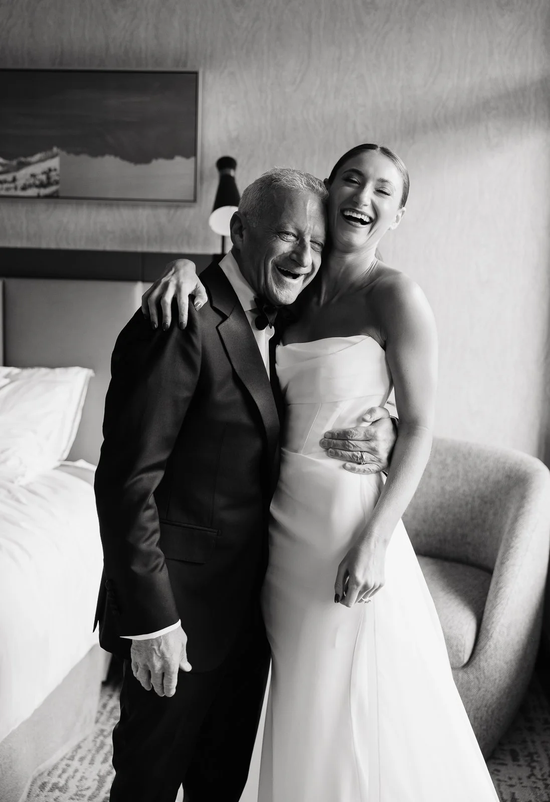A black and white photo of a man and woman laughing and hugging. The man is wearing a tuxedo, and the woman is in a strapless wedding dress in a hotel room.