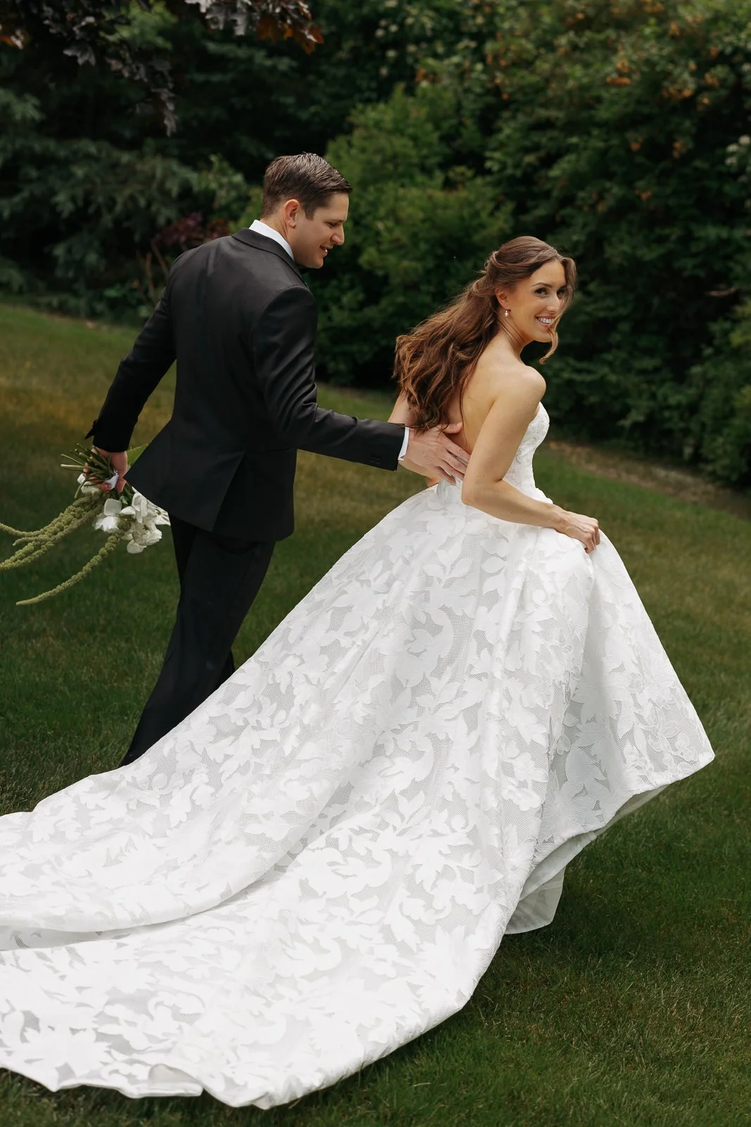 Bridalkind couple outdoors, woman in a white gown with a long train, man in a black tuxedo, green trees in the background.