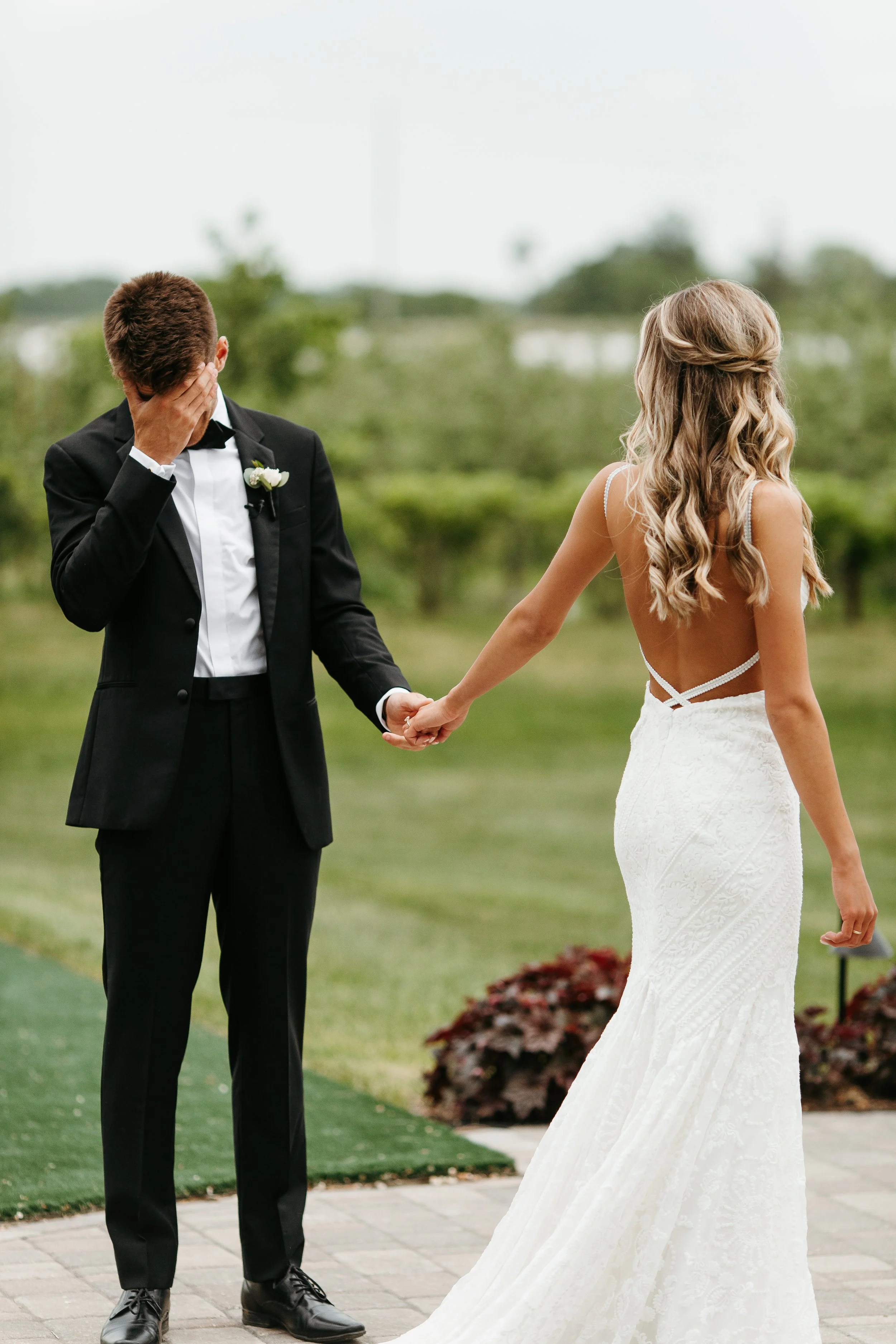 A bride and groom holding hands during an outdoor wedding ceremony. The groom, dressed in a black tuxedo, covers his face with his hand, while the bride, with long blonde hair styled in loose waves, is dressed in a white lace wedding gown with an ope