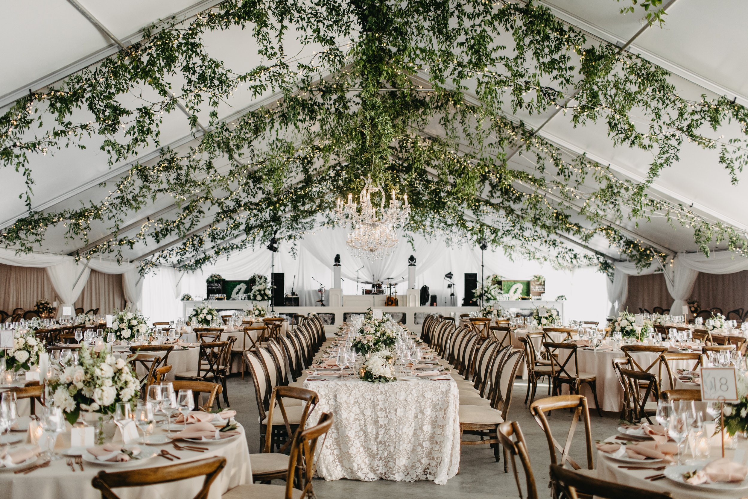 A decorated event tent with long banquet tables set for a wedding, featuring white tablecloths, floral centerpieces, and elegant glassware, with a chandelier hanging from the ceiling and string lights intertwined with greenery.