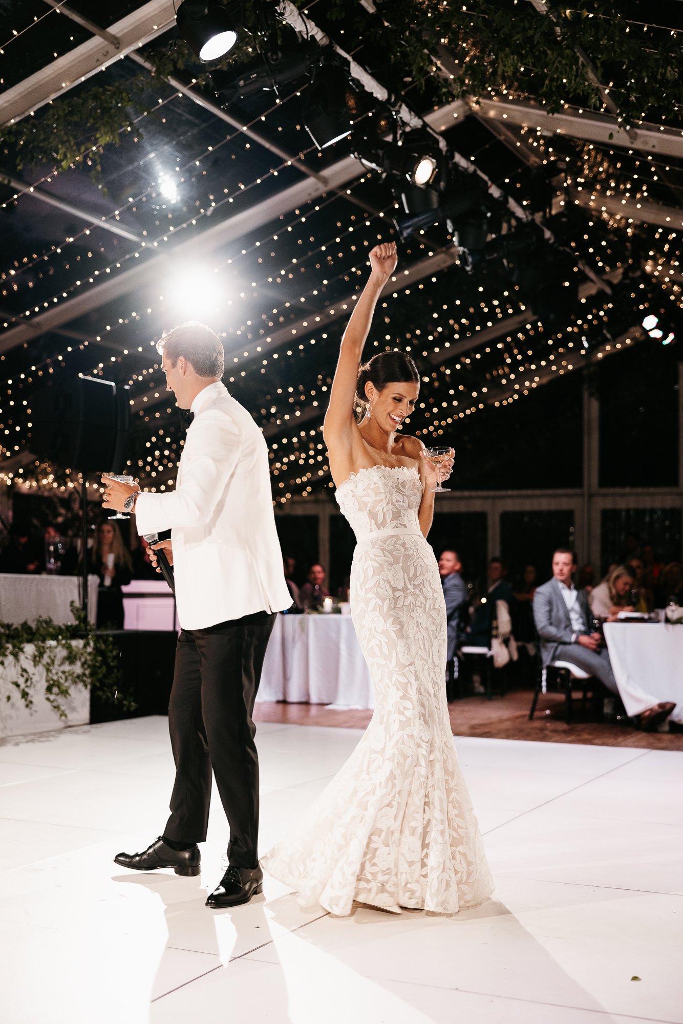 A woman in a white lace wedding dress dancing with a man in a white tuxedo jacket and black pants at a wedding reception under string lights.