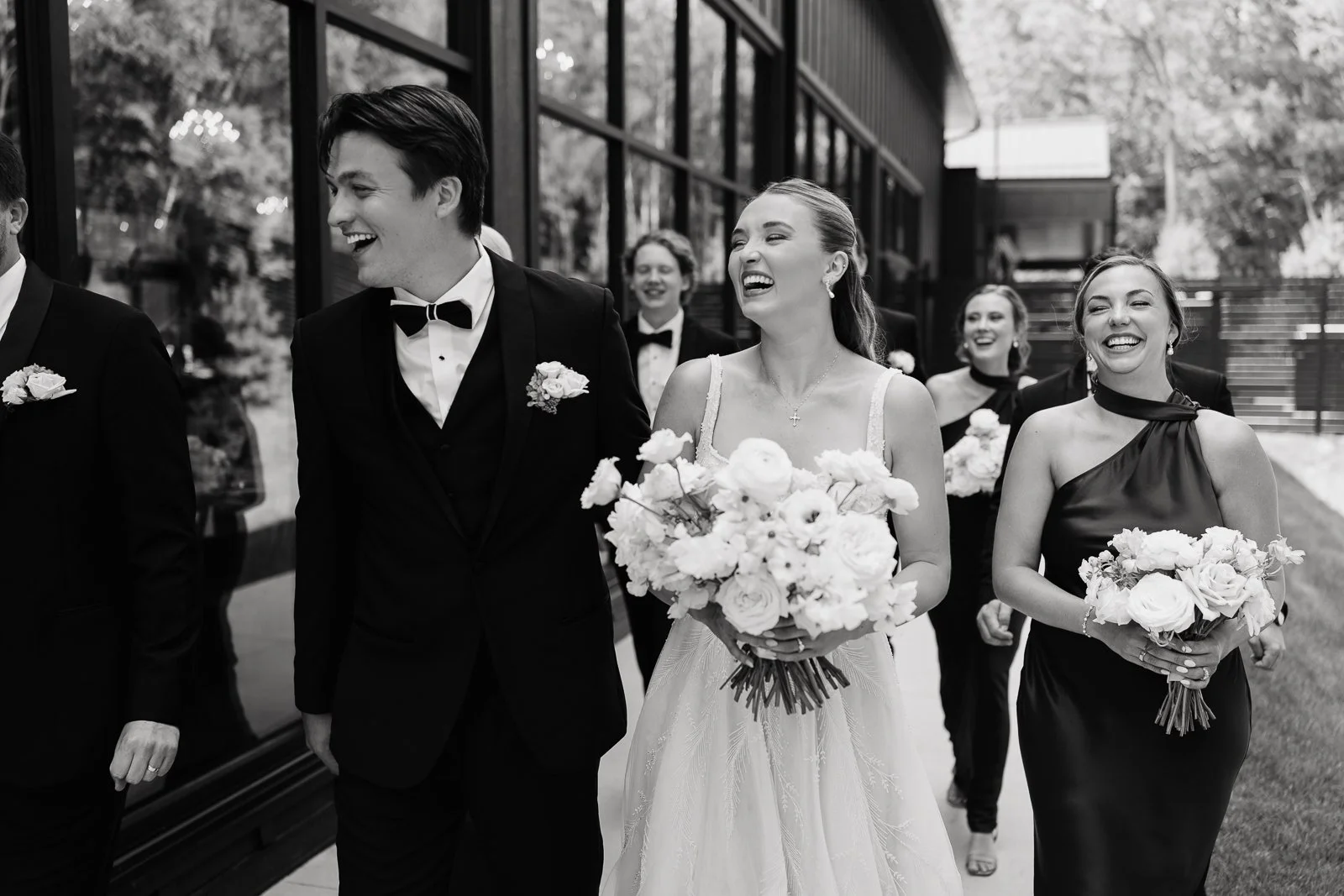 A wedding procession with the bride in a white dress and holding a bouquet, surrounded by bridesmaids and groomsmen in formal attire, walking outdoors near a building with reflective windows.
