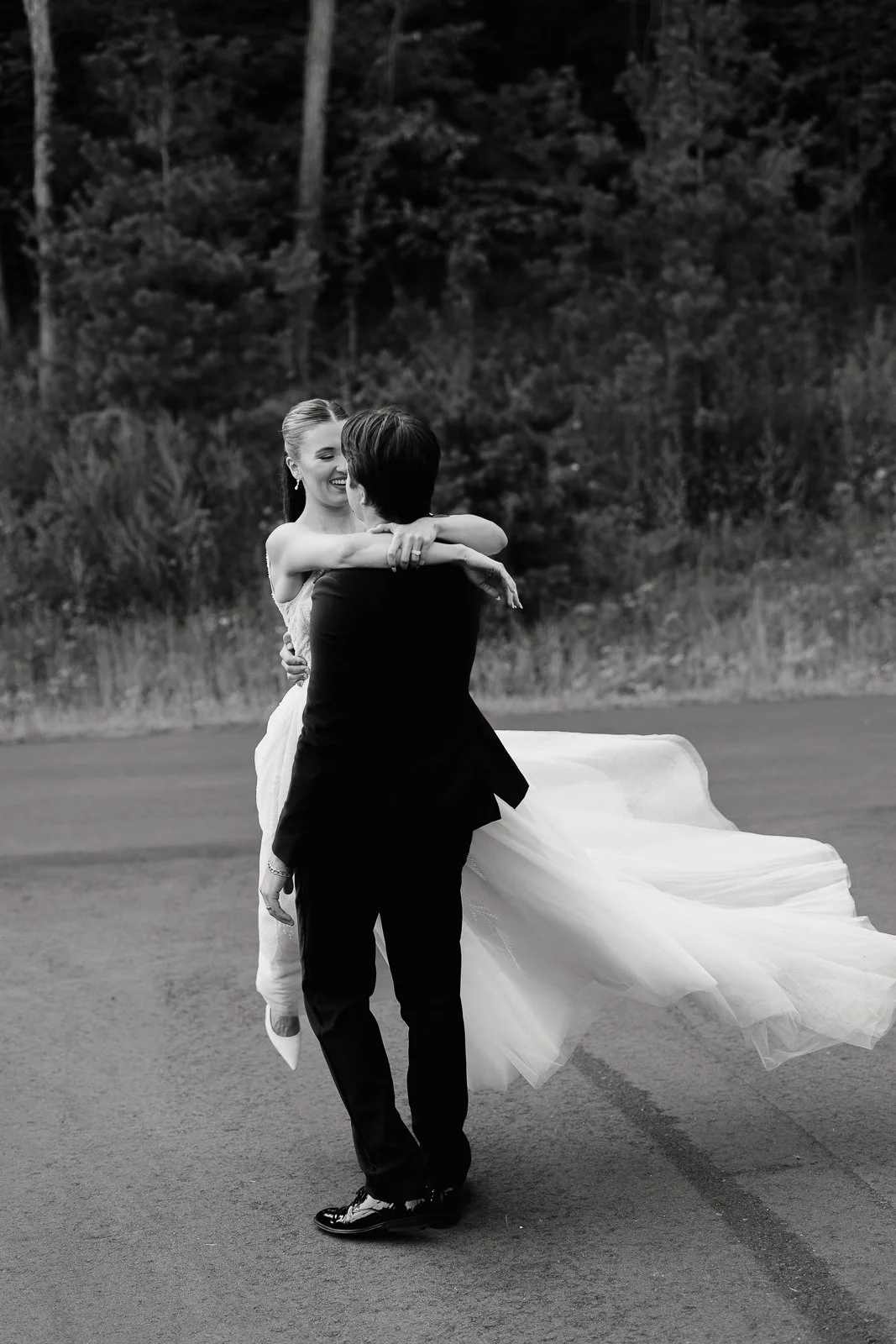 A black-and-white photo of a man and woman dancing outdoors on a road, with trees in the background. The woman is smiling wearing a wedding dress, and the man, in a suit, holds her as they dance.