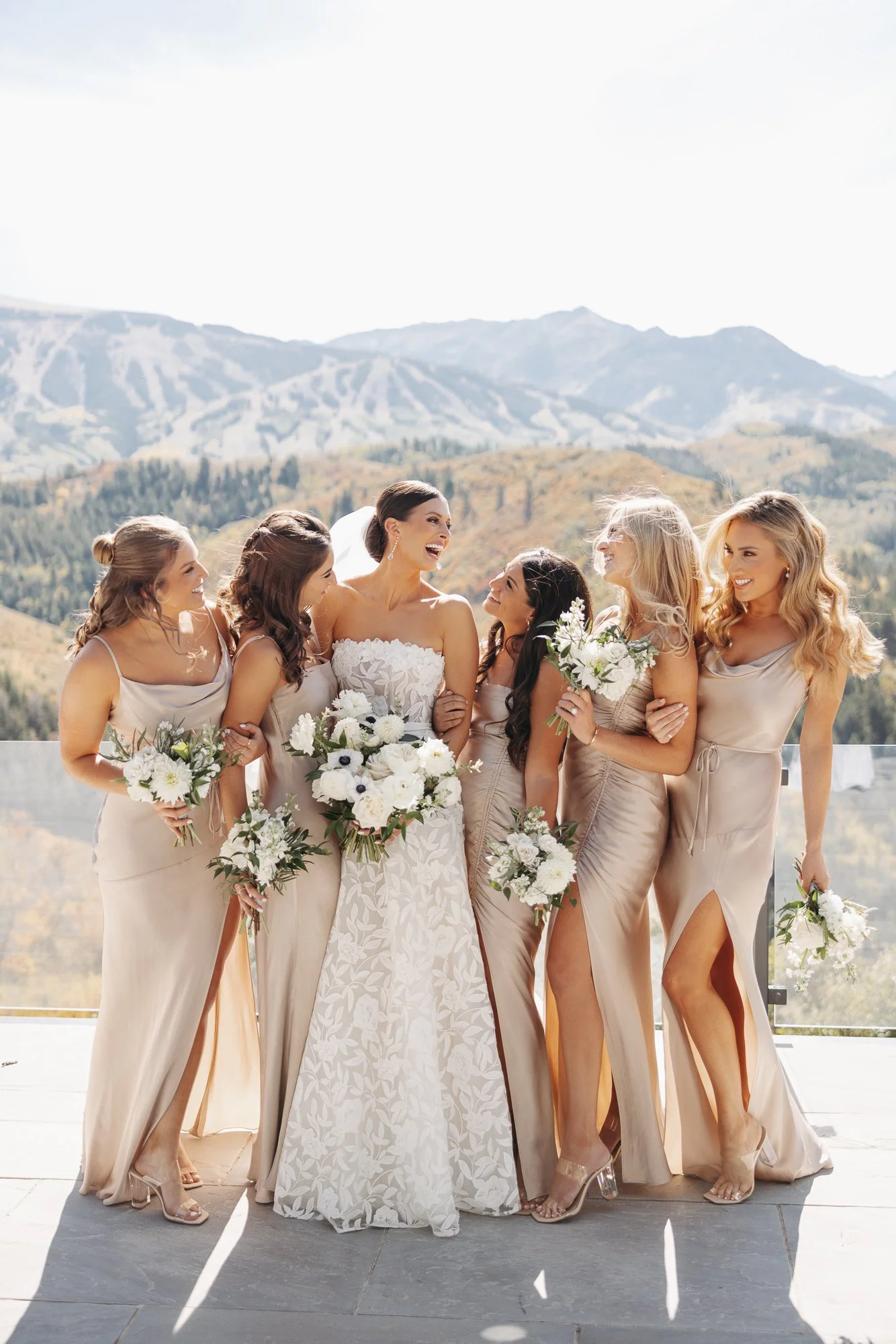 A bride and six bridesmaids standing together outdoors on a sunny day, with mountains in the background, all smiling and dressed in beige dresses, holding white and green floral bouquets.