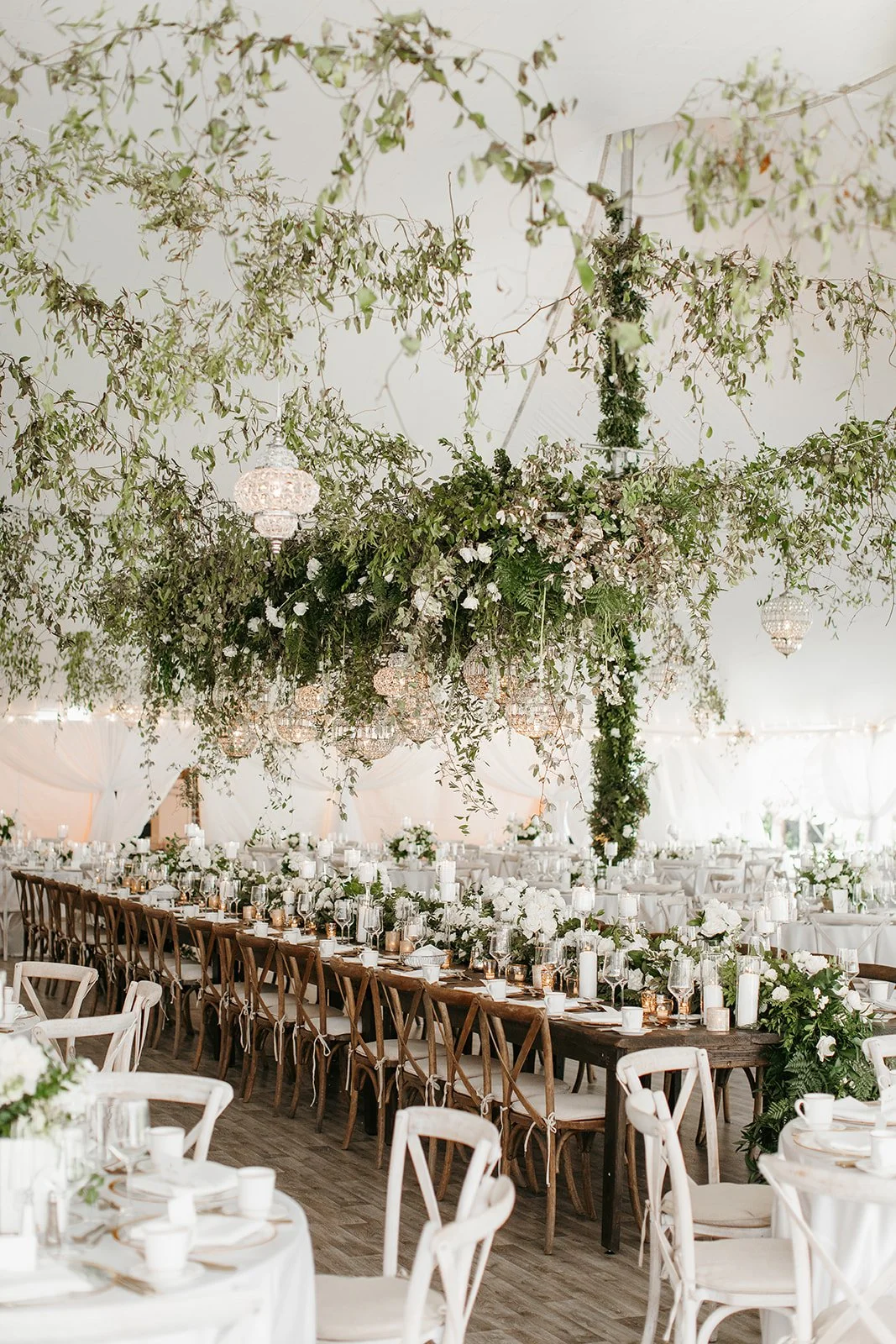 Elegant event reception with long dinner tables decorated with white floral arrangements, candles, and glassware, under hanging greenery and chandeliers.