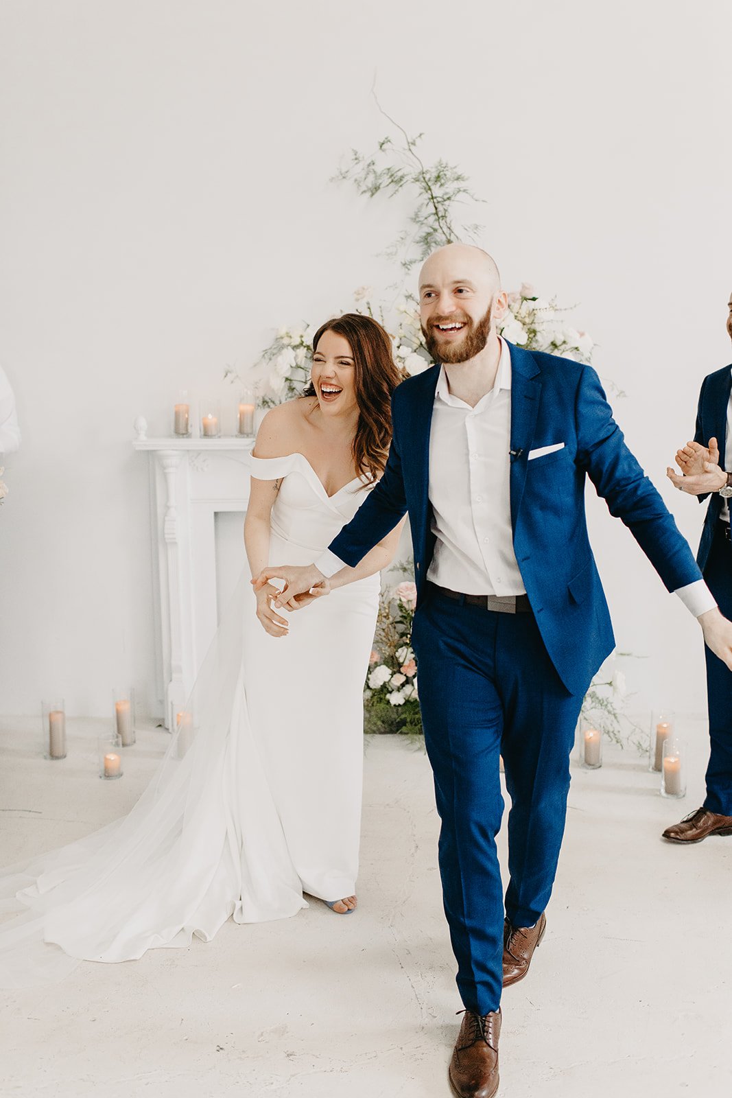 A bride and groom smiling and holding hands during their wedding ceremony, with candles and floral decorations in the background.