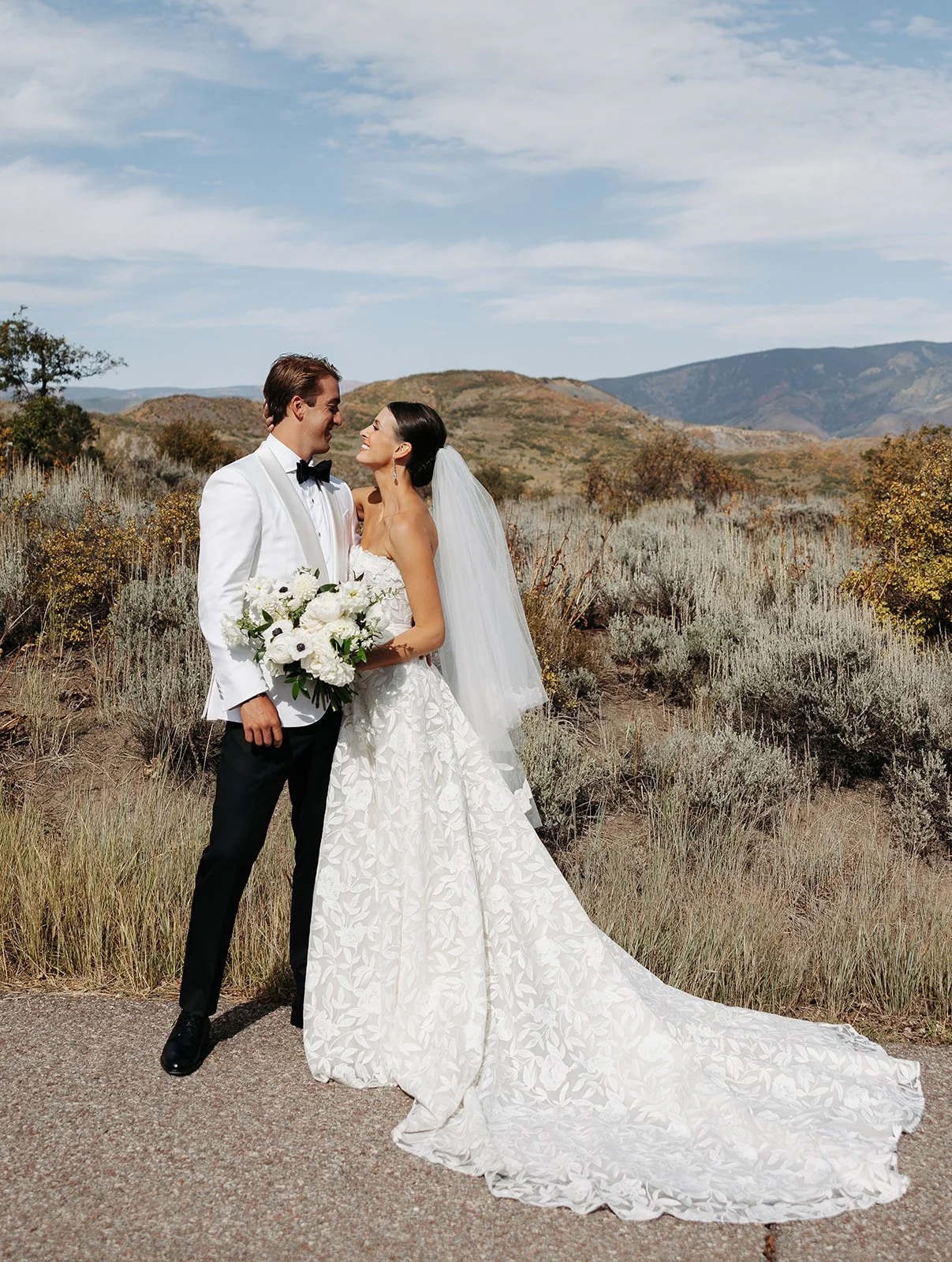 A bride and groom stand together outdoors in a dry, hilly landscape with shrubs and rolling hills in the background. The groom wears a white tuxedo jacket and black pants, and the bride wears a strapless white wedding gown with a long train and veil.
