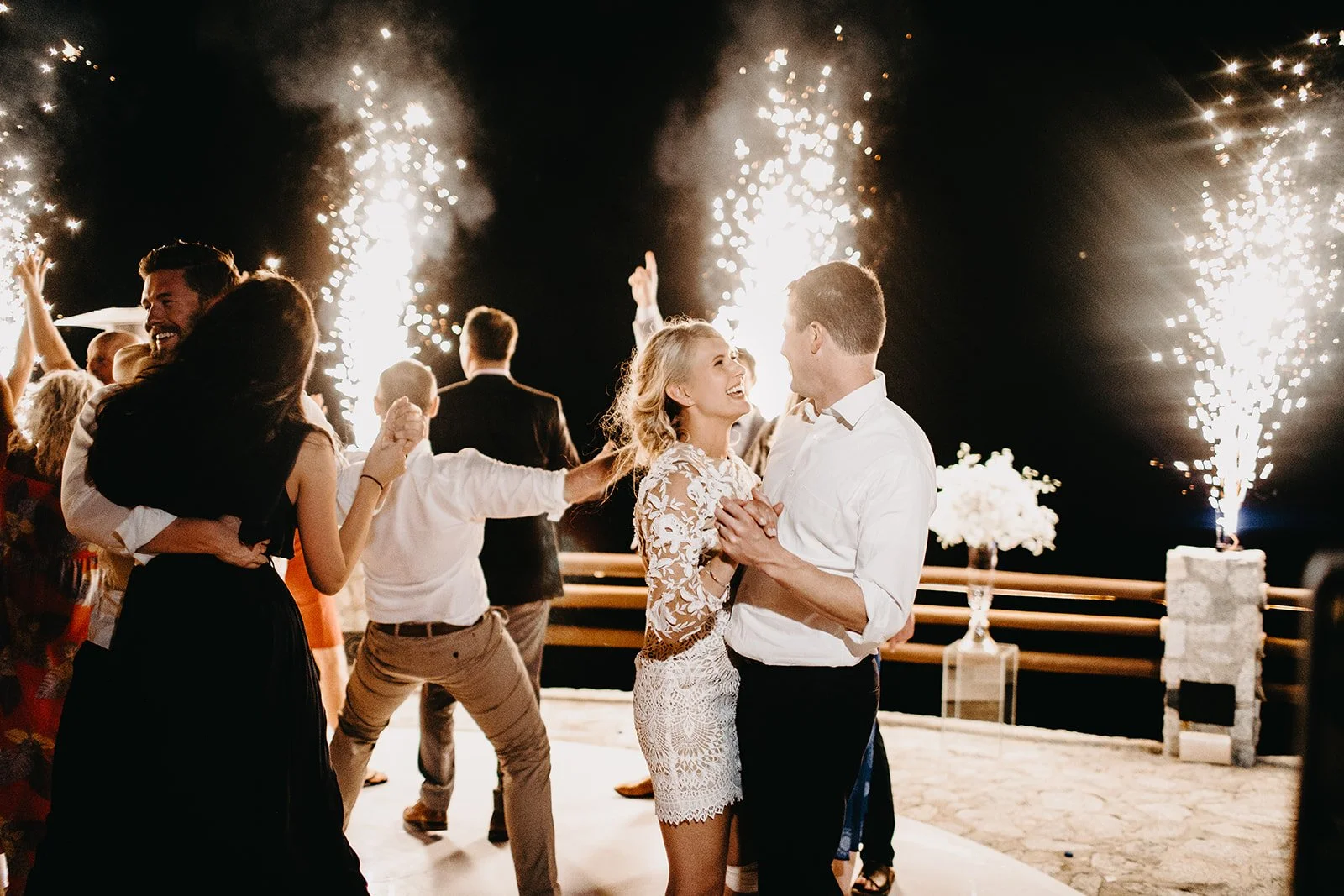People dancing and celebrating at a nighttime wedding reception with fireworks in the background.