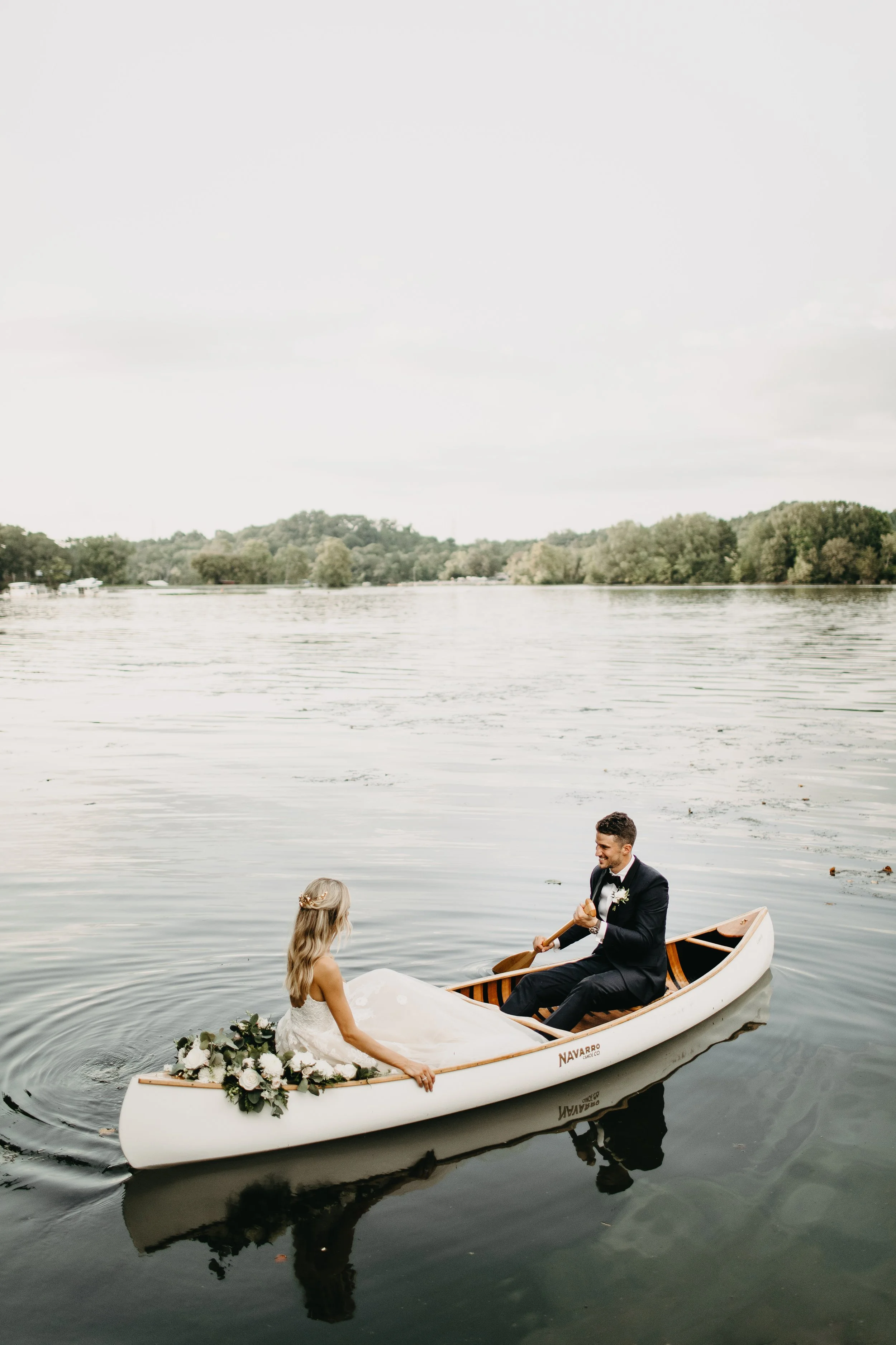 A bride and groom in a boat on a calm lake during sunset, with lush trees in the background. The bride is holding a bouquet of flowers, and the groom is rowing the boat. They are dressed in wedding attire, celebrating their wedding.