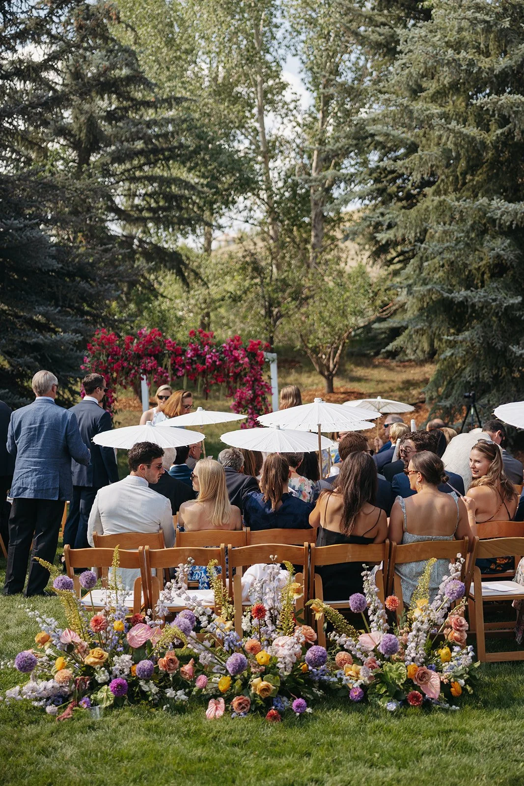 An outdoor wedding ceremony with guests seated on wooden chairs, some holding umbrellas, in a garden setting with tall trees and pink flowering arch in the background. Colorful flowers are arranged in front of the seating area.