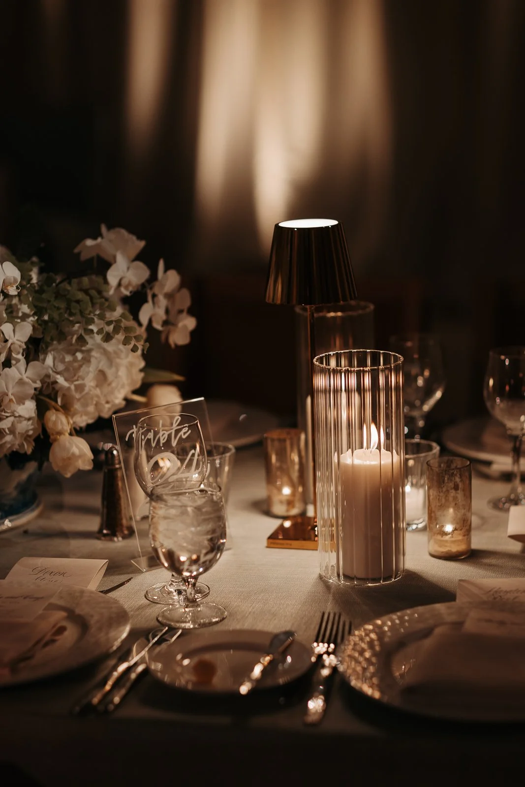 Elegant dining table with candles, flowers, and glassware, illuminated by warm candlelight and a small lamp, set for a formal event or celebration.