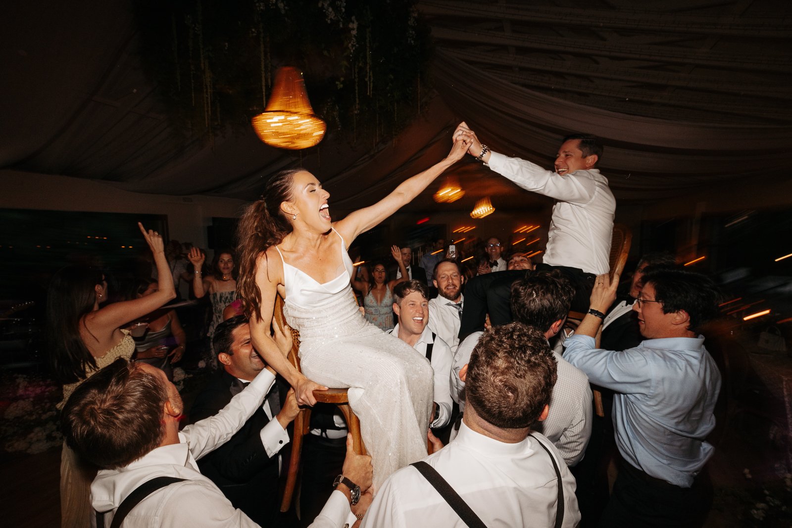 A lively celebration with a group of people lifting a woman and man in formal attire, possibly at a wedding or party, inside a decorated venue with warm lighting.
