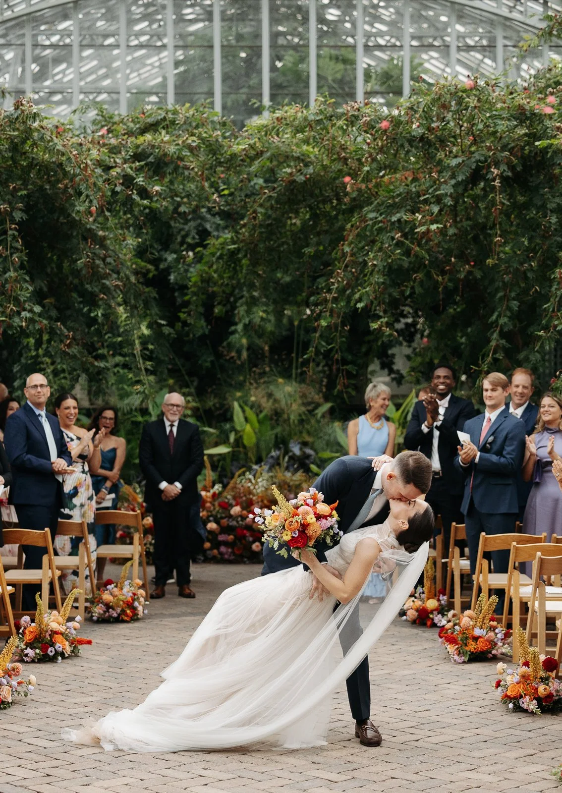 A wedding ceremony taking place inside a greenhouse, with the bride and groom sharing a kiss, surrounded by guests clapping and smiling, with lush green plants and flowers.