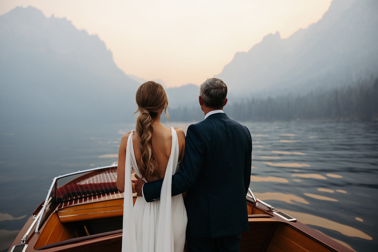 A couple in wedding attire on a boat, looking at a misty mountain landscape across a lake during sunset.