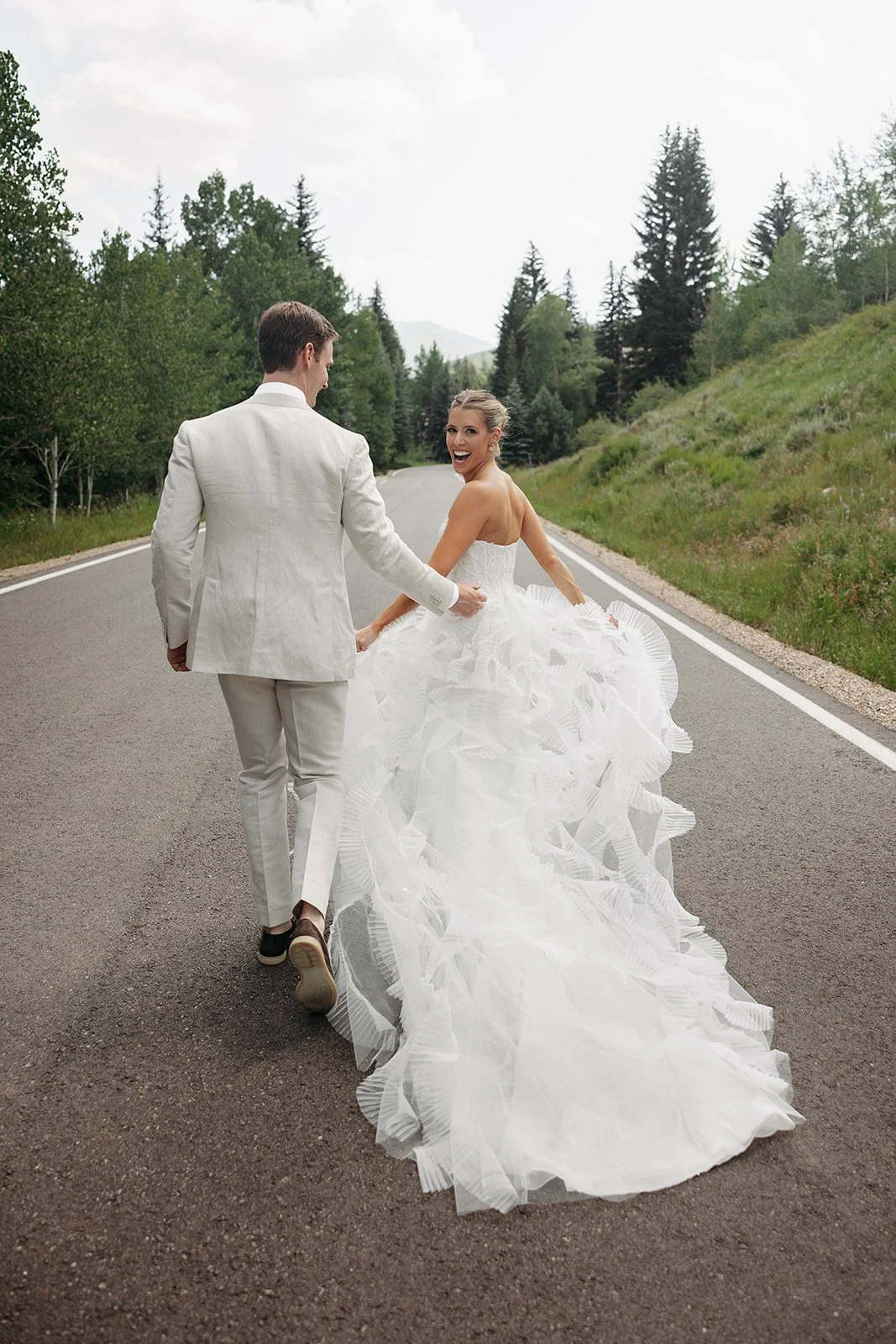 A bride and groom walking hand in hand on an empty road surrounded by lush greenery, with the bride smiling and looking back.