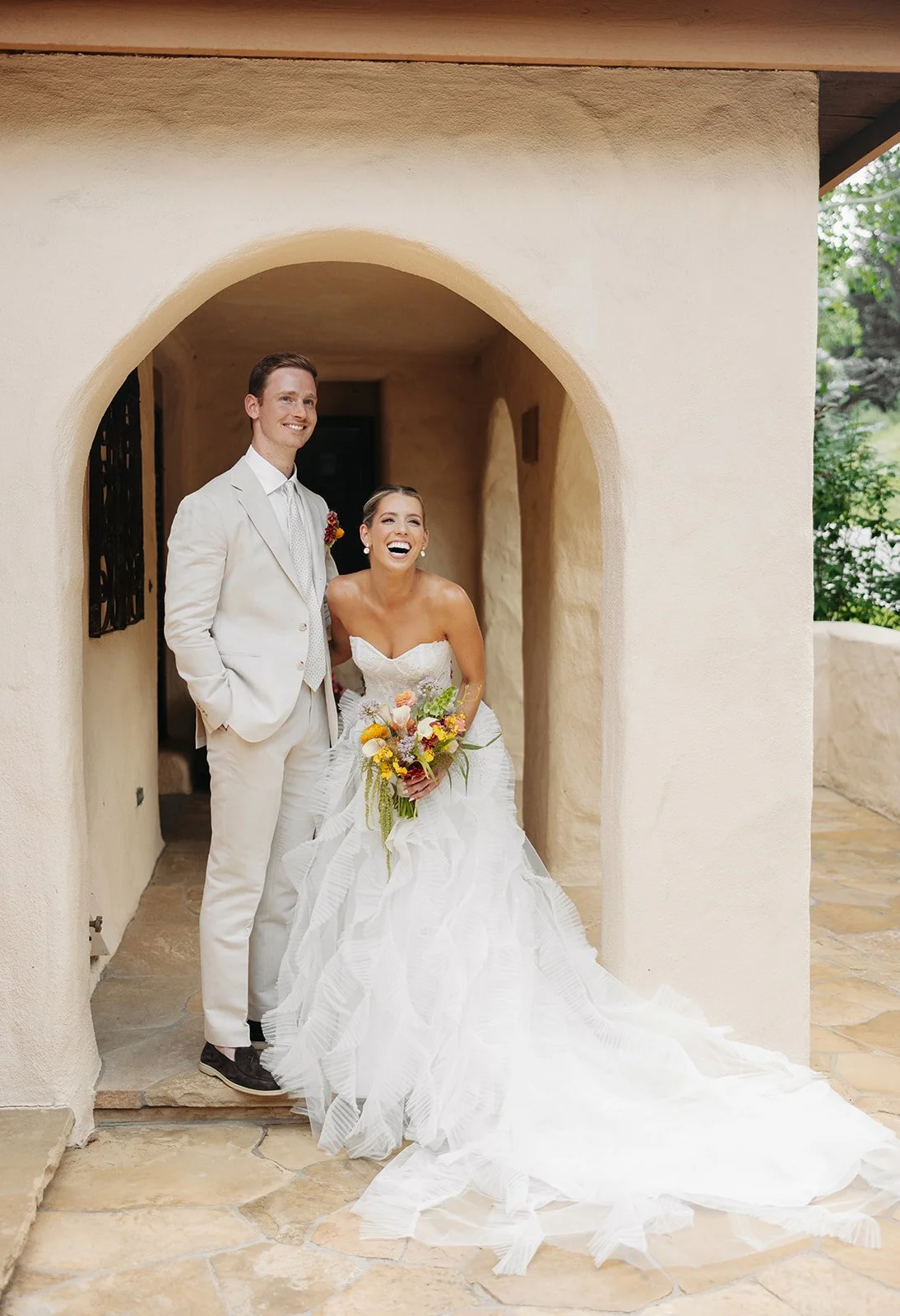 A happily married couple, the bride in a white wedding gown holding a bouquet of colorful flowers and the groom in a white suit, standing under an archway with beige stucco walls, during their wedding day.
