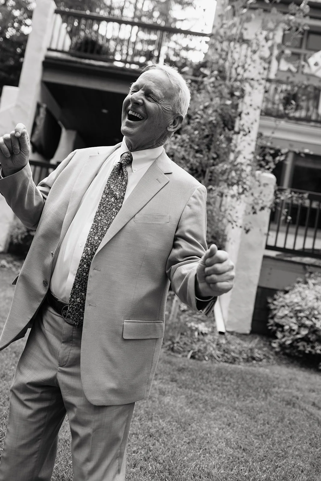 An elderly man in a light suit and patterned tie, smiling joyfully, dancing outdoors near a garden and residential building, in black and white.