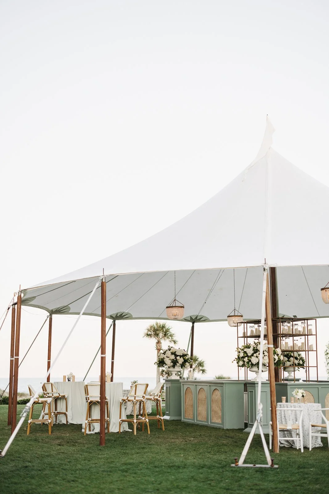 Outdoor wedding reception setup under a large white tent with tables, chairs, floral arrangements, and lanterns on a grassy lawn.