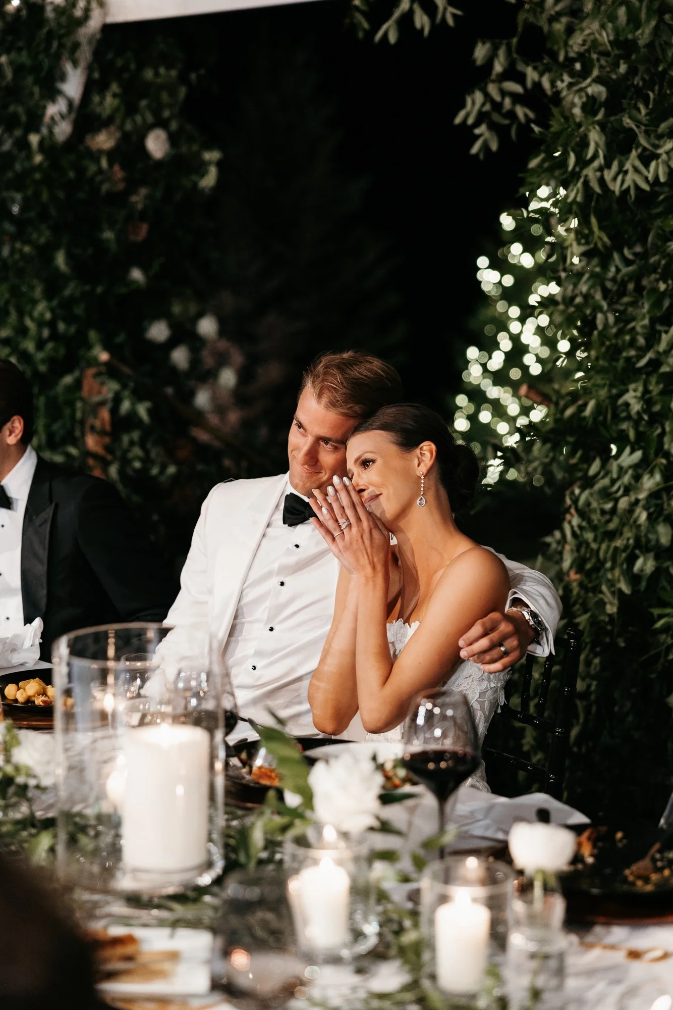 A couple dressed in formal wedding attire sitting close together at a wedding reception table, with the woman resting her hand on her cheek and showing her wedding rings. The setting is decorated with candles and greenery, with blurred string lights 