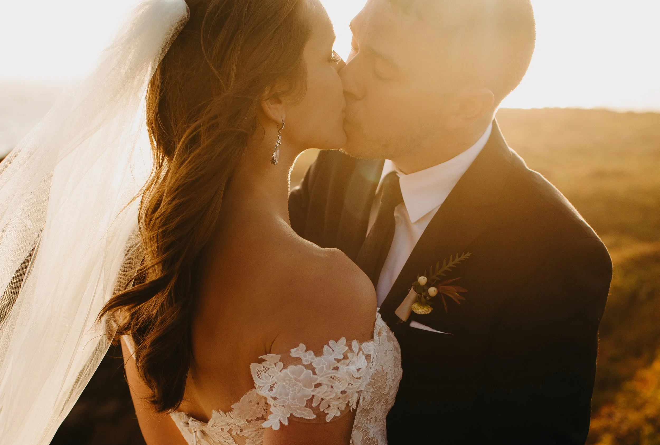 A bride and groom kissing outdoors during sunset, with the bride wearing a white lace wedding dress and veil, and the groom in a black tuxedo with a boutonniere, emphasizing a romantic moment.