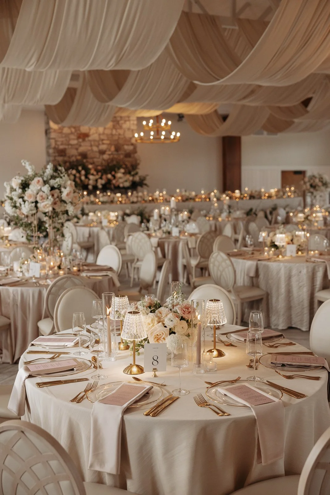 Elegant wedding reception decorated with cream-colored tablecloths, floral centerpieces, and gold accents. Soft lighting and draped fabric ceiling enhance the sophisticated atmosphere.
