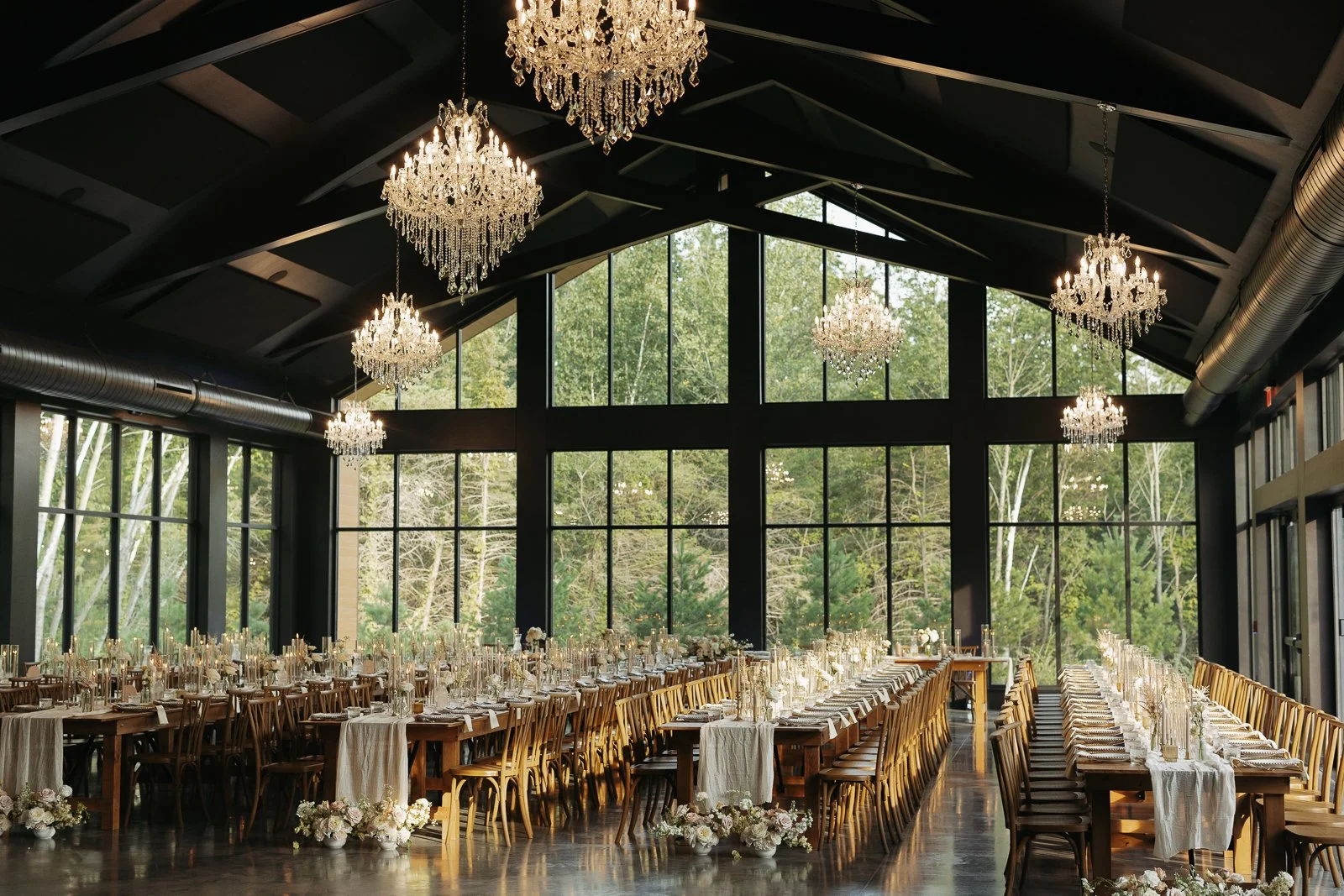 Elegant event space with large floor-to-ceiling windows, crystal chandeliers hanging from the dark ceiling, and a long dining table set with white and gold accents, surrounded by wooden chairs, decorated with floral arrangements at the end, overlooki