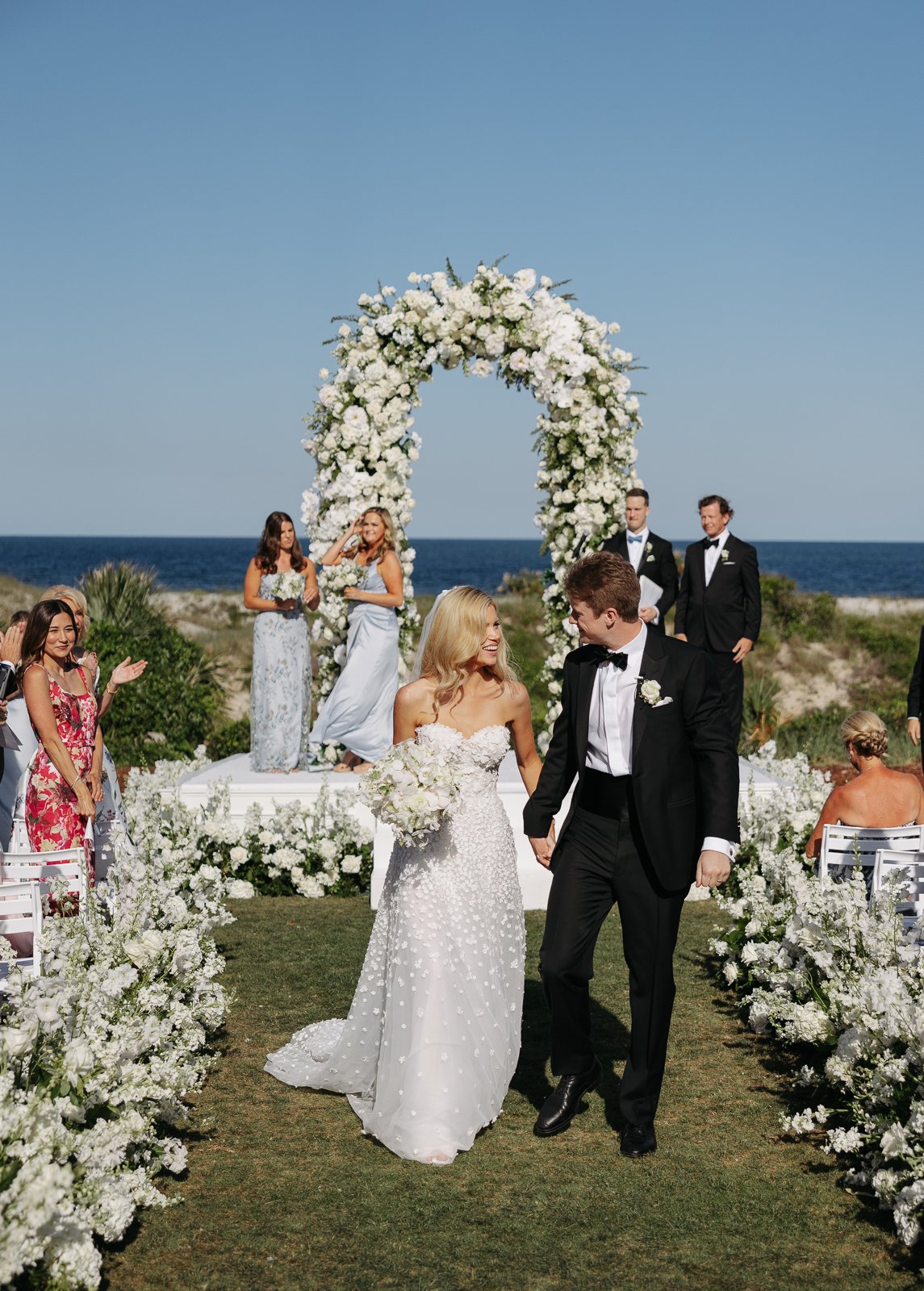 A wedding ceremony taking place outdoors on a sunny day by the beach, with a floral arch and the bride and groom walking hand in hand.