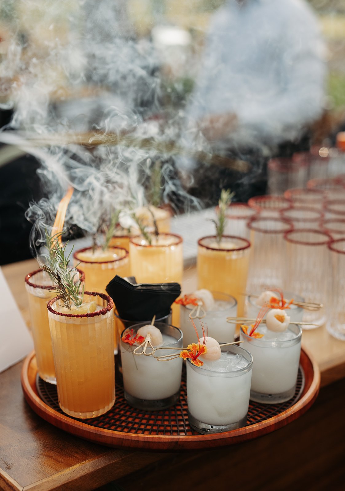 Several glasses with smoked drinks, garnished with herbs and flowers, are arranged on a round wooden tray.