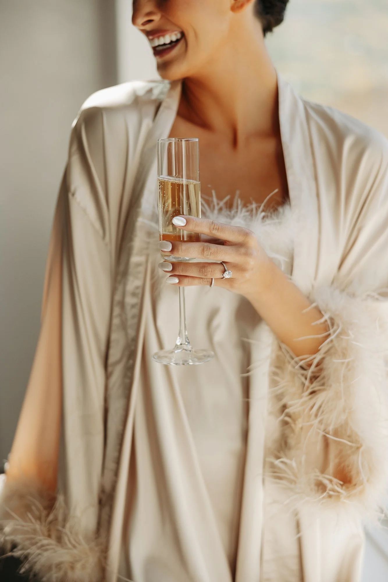 Woman holding a glass of champagne, wearing an elegant cream-colored satin dress and a ring, smiling and laughing.