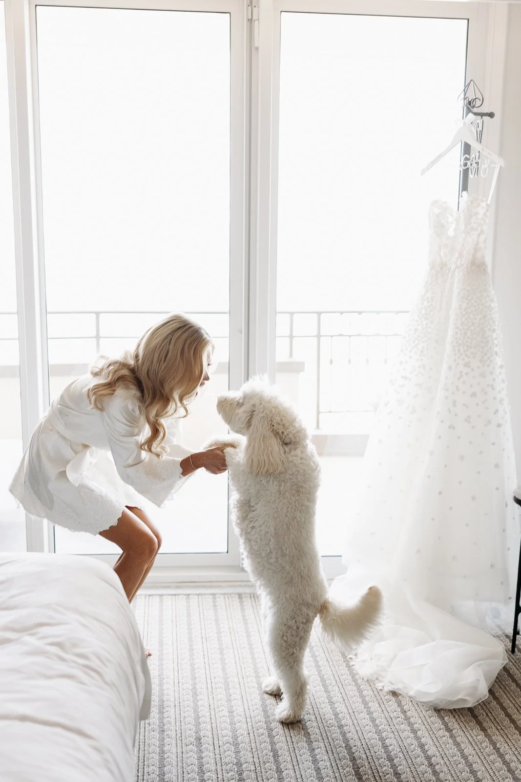 A woman in white dress bent down and holding hands with a large fluffy white dog inside a bright room near a balcony door, with a wedding dress hanging on a stand nearby.