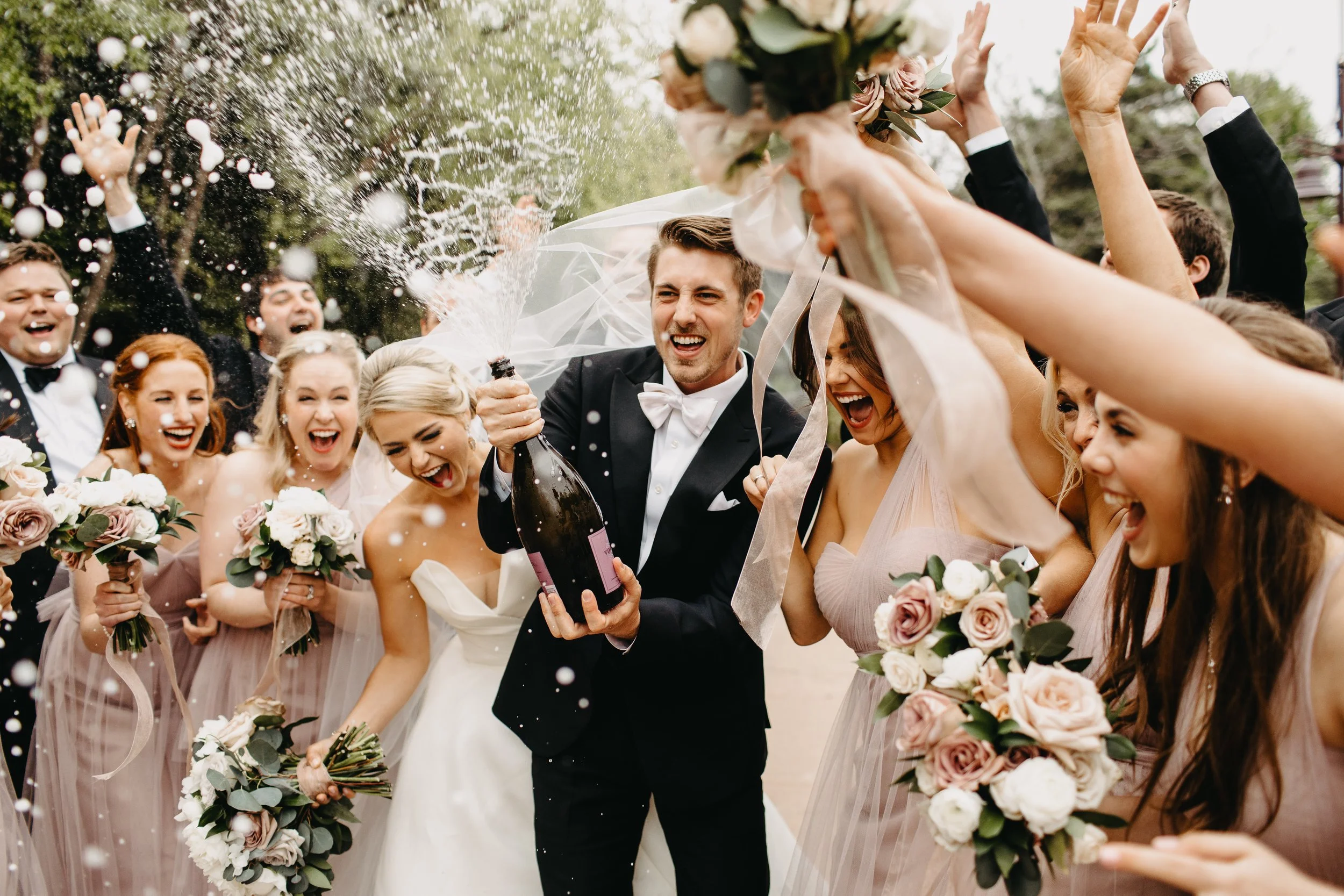 Bride and groom celebrating with wedding guests, some holding bouquets, in outdoor setting, with champagne