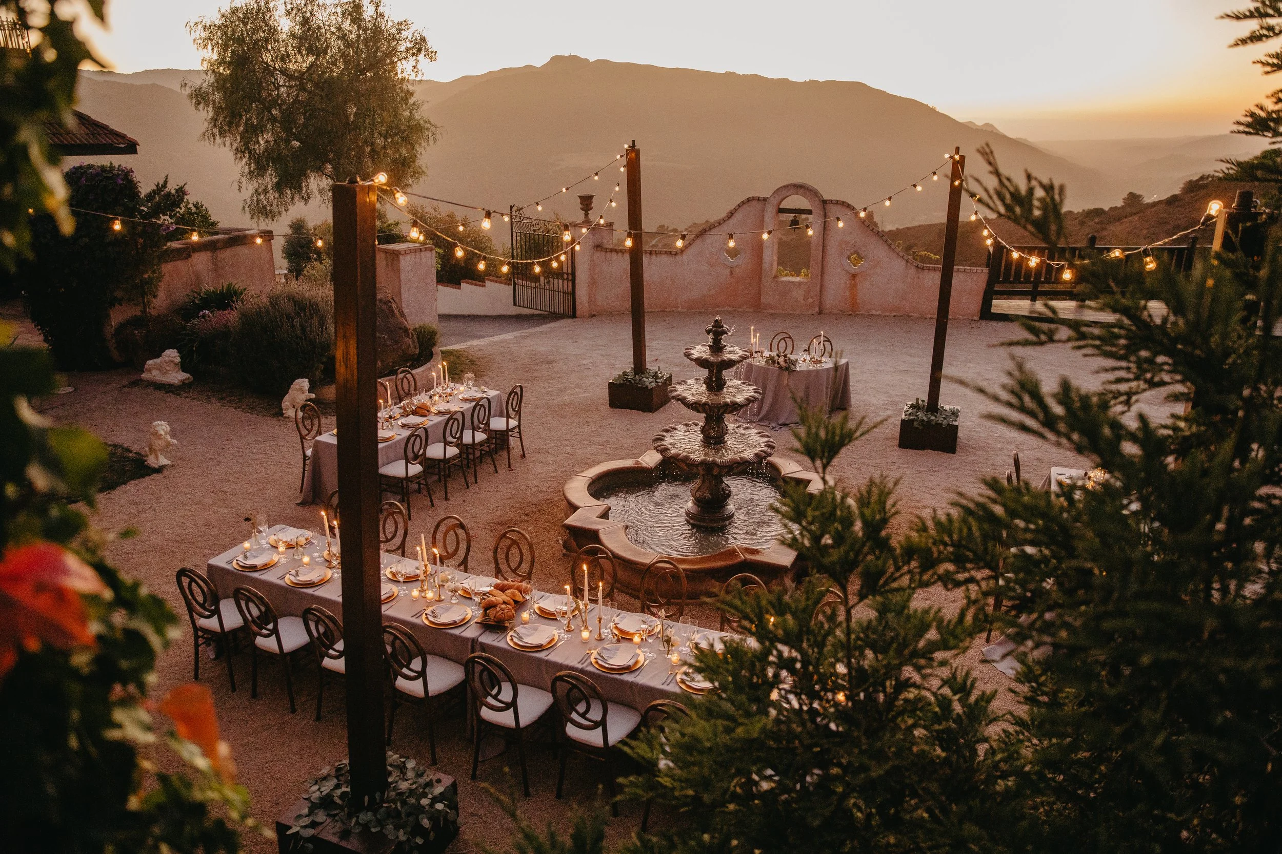 An outdoor dining area during sunset with a long table set with plates, cutlery, candles, and bread, surrounded by string lights, a fountain, plants, and mountains in the background.