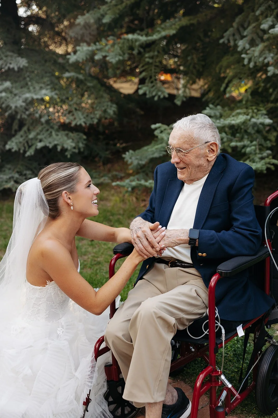 A young woman in a wedding dress smiling and holding hands with an elderly man in a wheelchair, outdoors with pine trees in the background.
