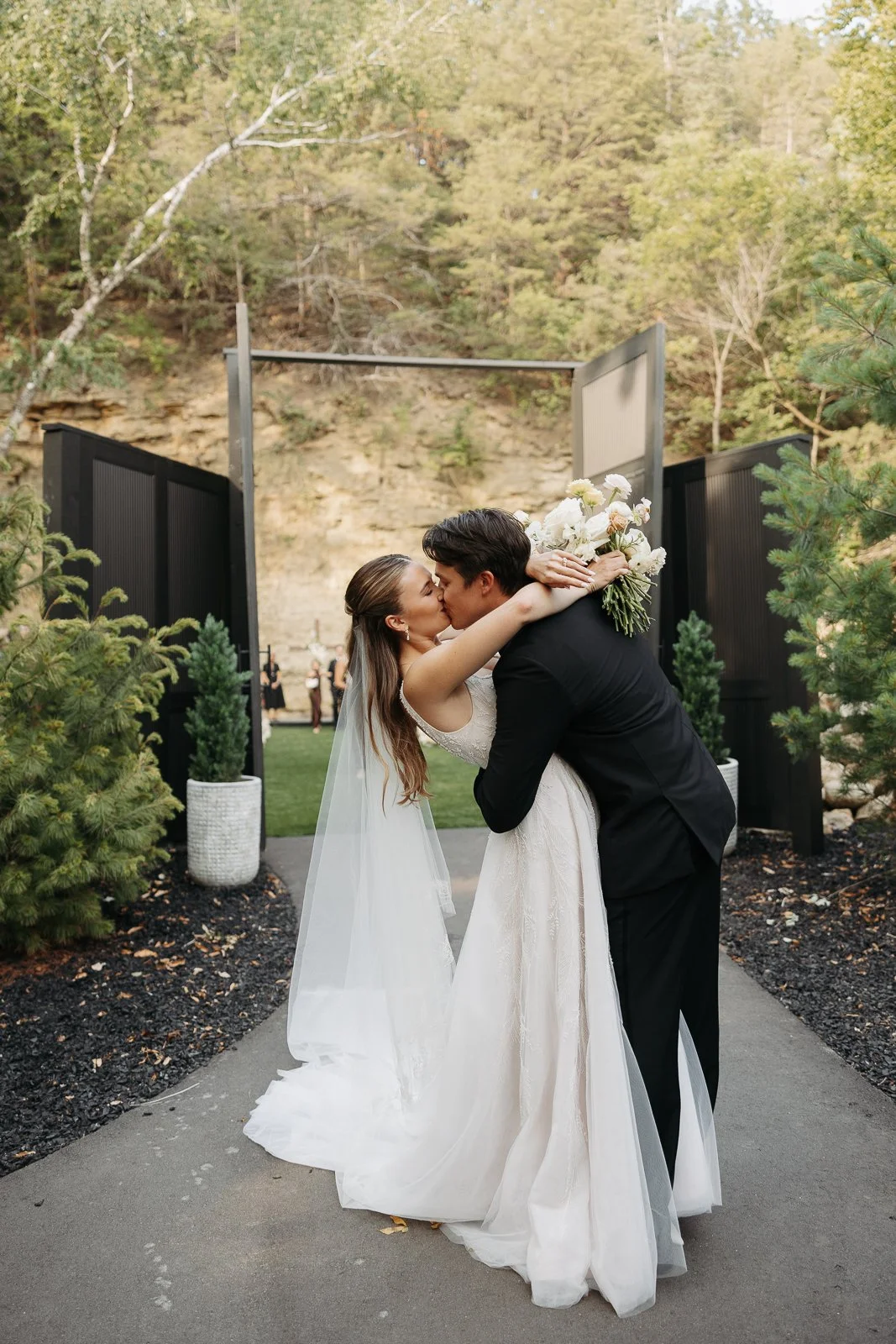 A bride and groom sharing a kiss outdoors during their wedding, with the bride holding a bouquet of white flowers and wearing a white wedding dress, and the groom in a black suit, surrounded by trees and black gates.