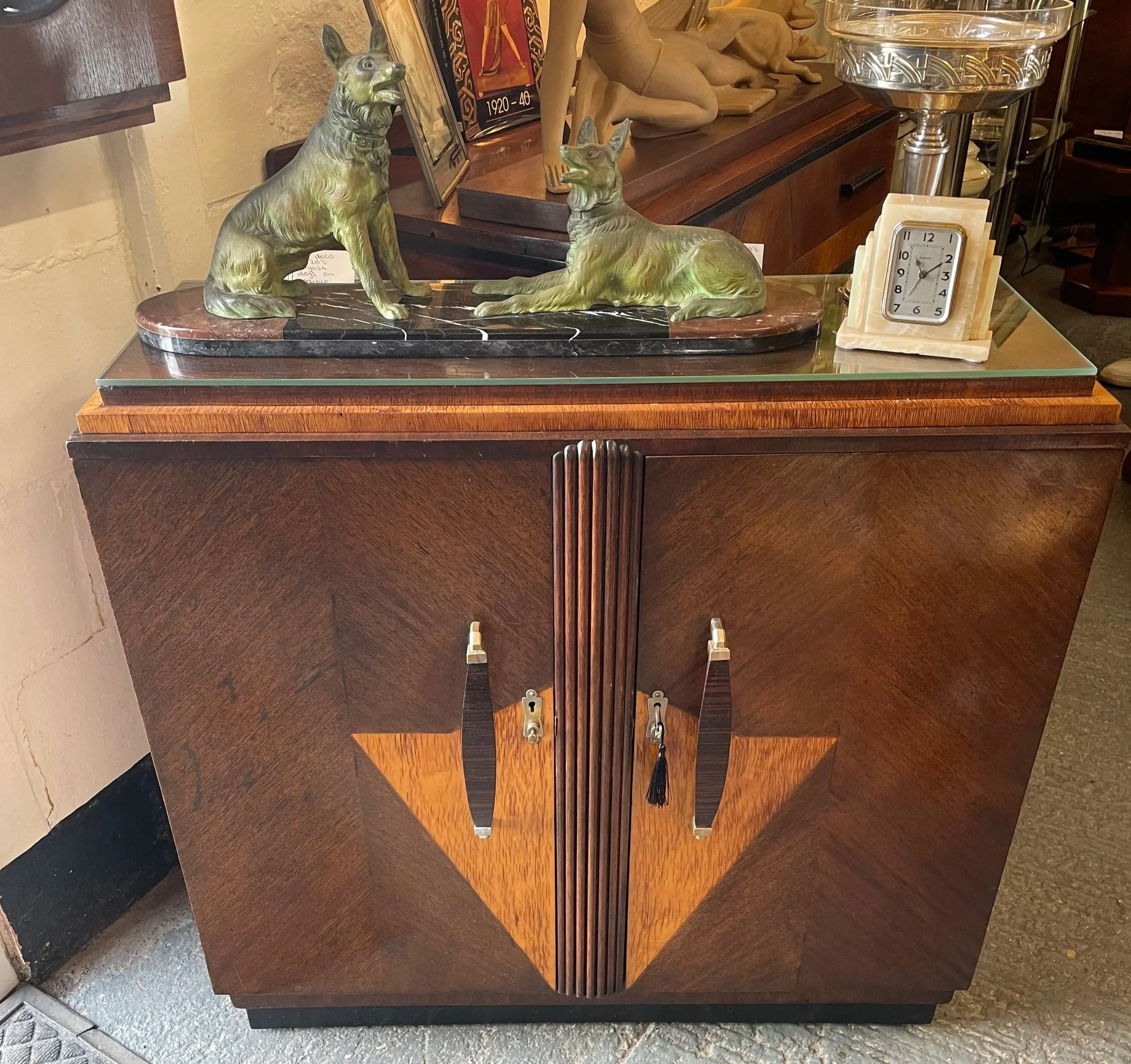 Decorative wooden dresser with brass lock, figurines of seated and lying dogs, a clock, and various artwork and figurines on top.
