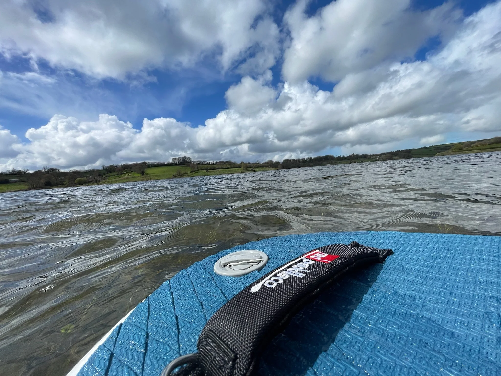 View from a paddleboard on a body of water with a wrist leash attached, green hills with trees and fields in the distance under a partly cloudy sky.