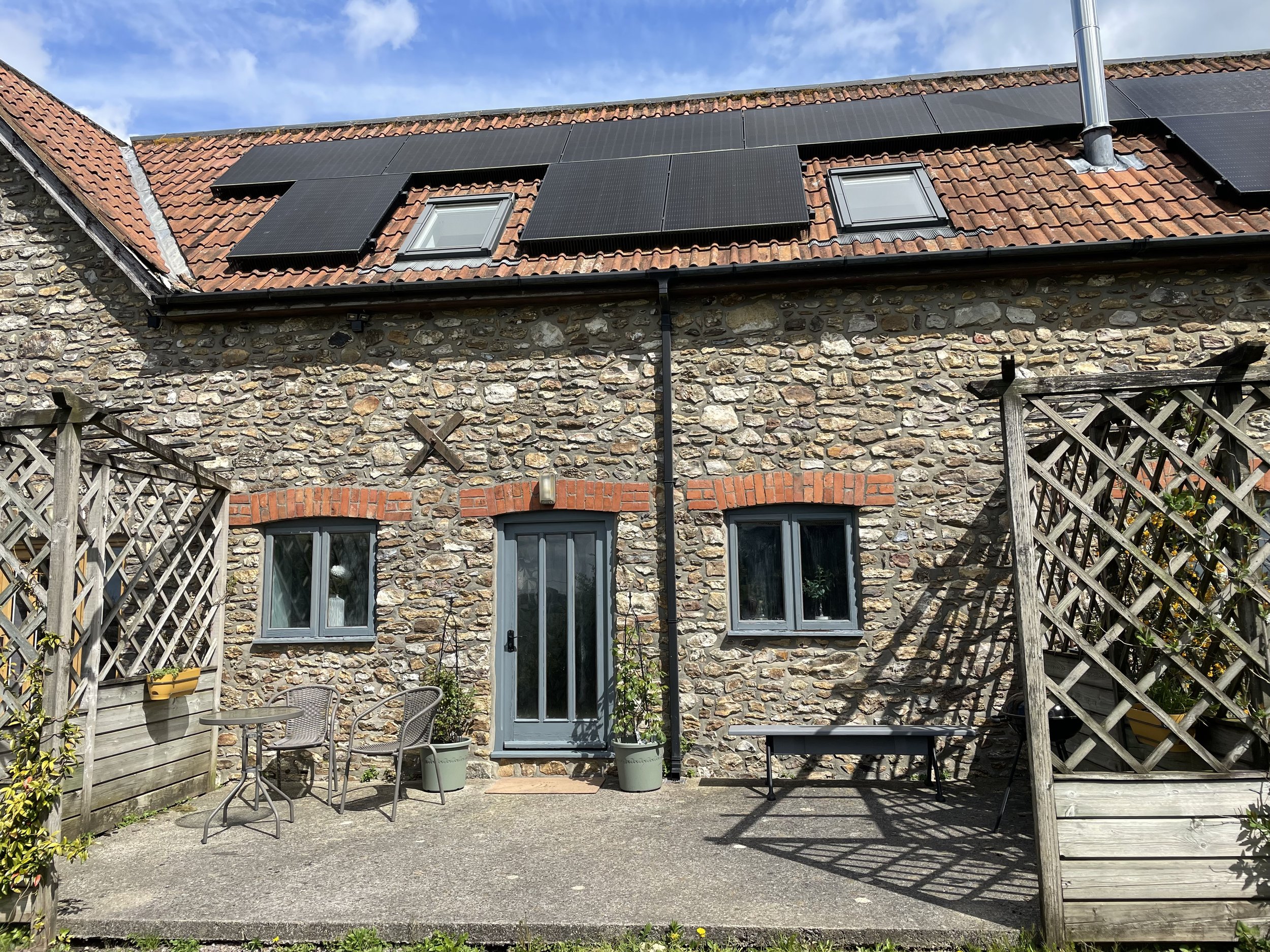 Backyard patio with a stone house wall, two skylight windows, and solar panels installed on the roof. There is outdoor furniture including a round table with chairs, and wooden trellises with plants growing on them.