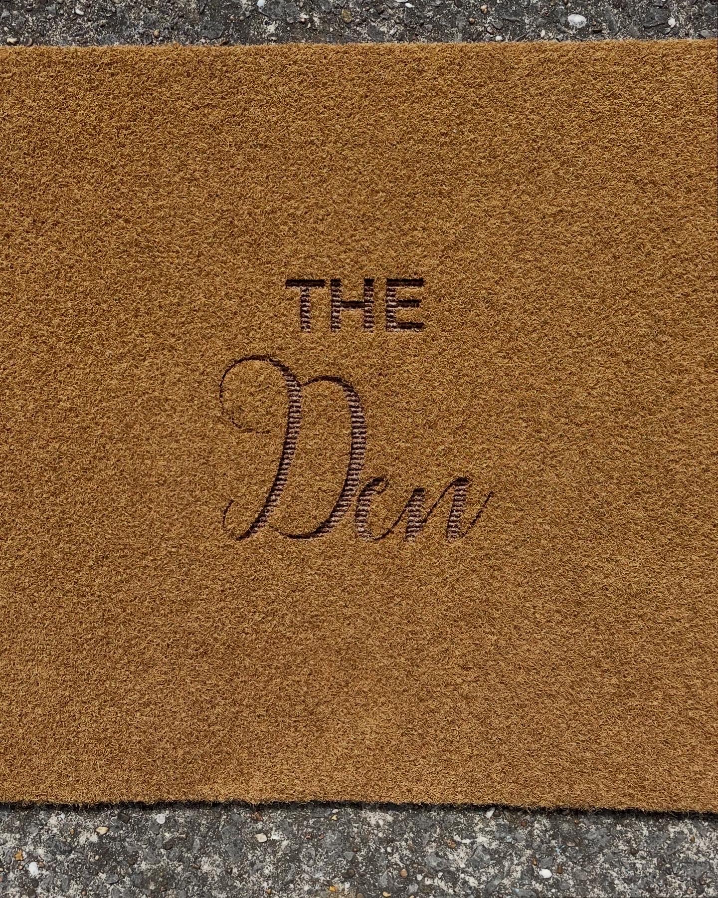 Close-up of a door mat with black stitched text reading "The Den".