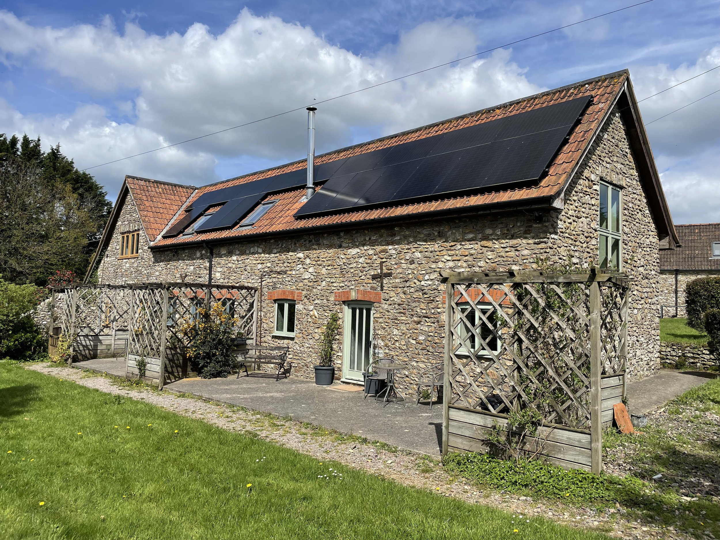 A stone house with red-tiled roof equipped with black solar panels. There is a small yard with garden furniture, a wooden fence, and a green lawn, under a partly cloudy sky.