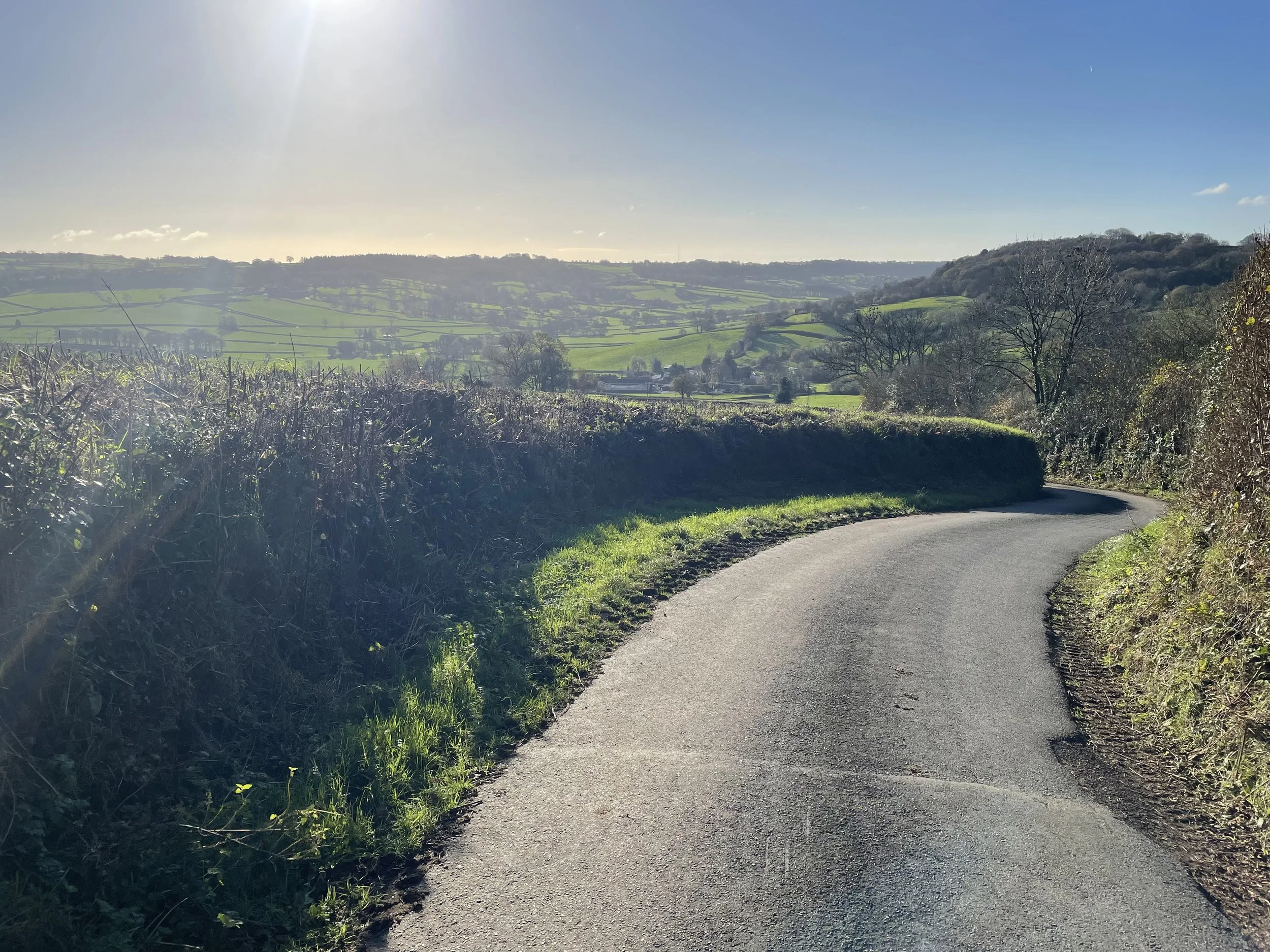 A winding country road runs through rolling green hills with trees and fields, under a mostly clear blue sky with the sun shining.
