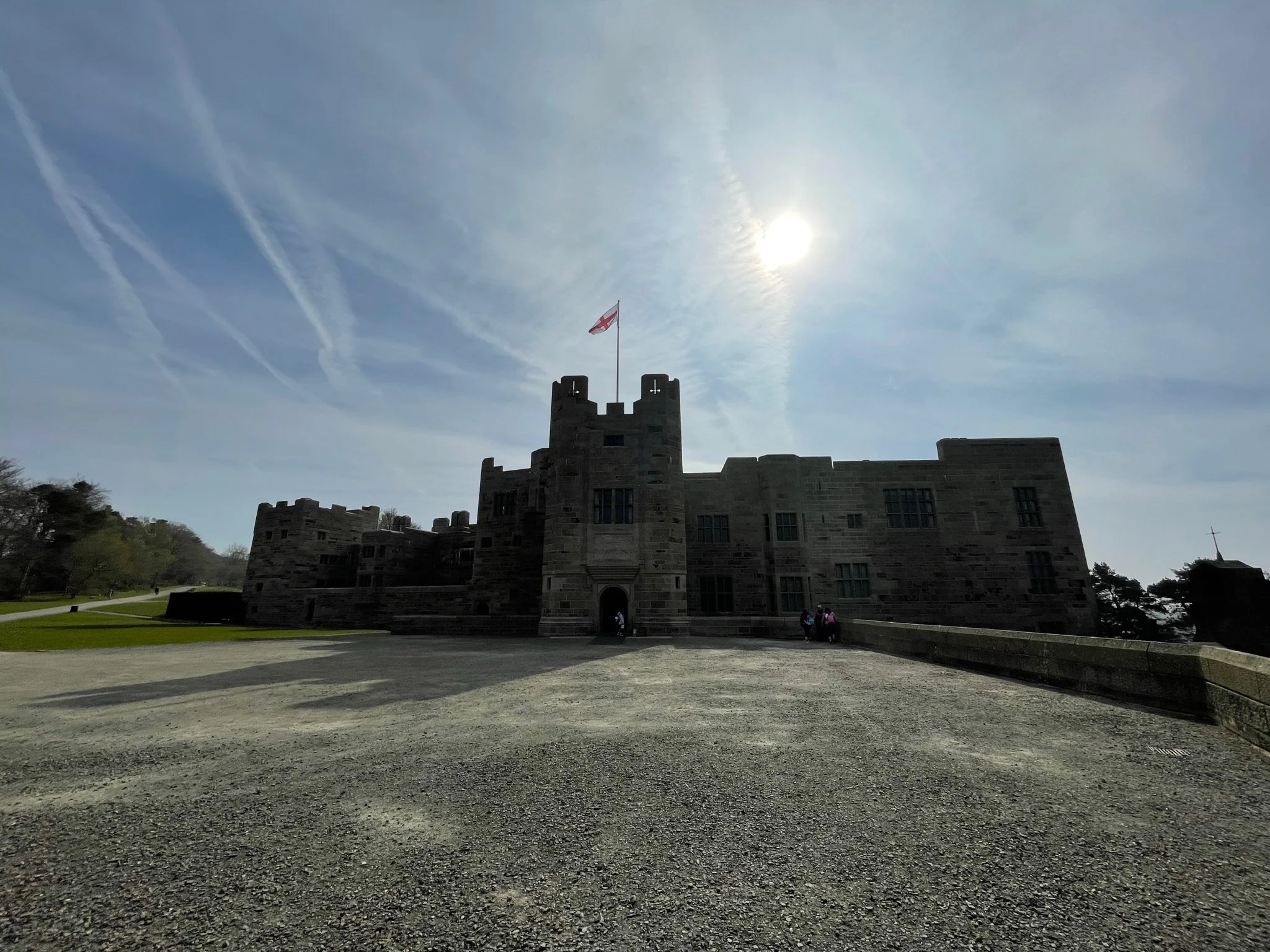 A historic stone castle with a flag on top, set against a bright, partly cloudy sky with the sun shining through. There are a few visitors near the entrance, and a large open gravel area in the foreground.