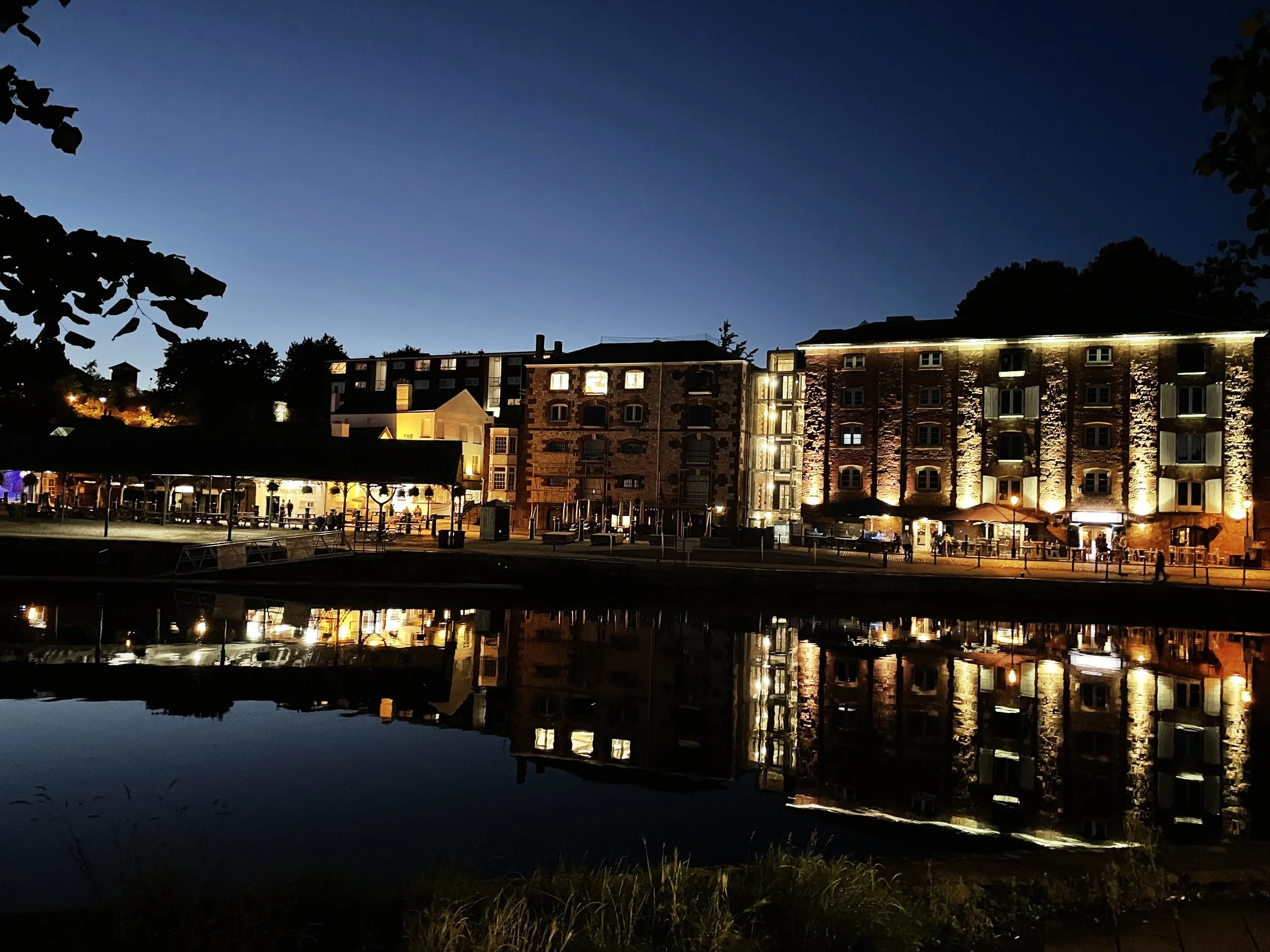 Nighttime view of buildings illuminated with lights, reflected on a calm body of water, with trees and a dark sky overhead.