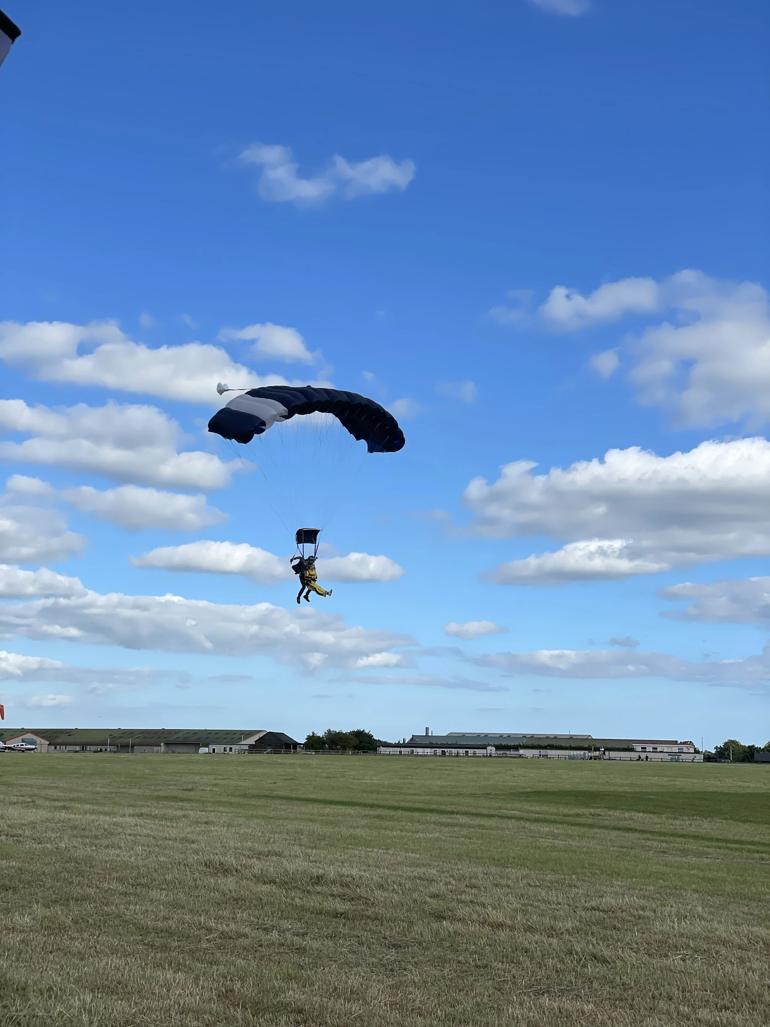 A person tandem skydiving with a black and white parachute over a flat grassy field with a few buildings in the distance and a blue sky with scattered clouds.