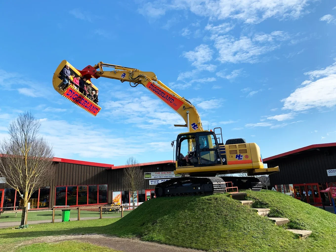 A large yellow excavator with a circular arm that extends upward, lifting a ride labeled 'DIGGERLAND' with children sitting inside, at an amusement park or playground.