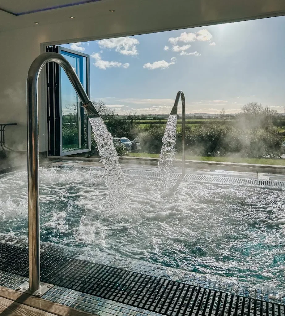 Indoor hot tub with open window showing a view of a cloudy sky, trees, and green fields outside.