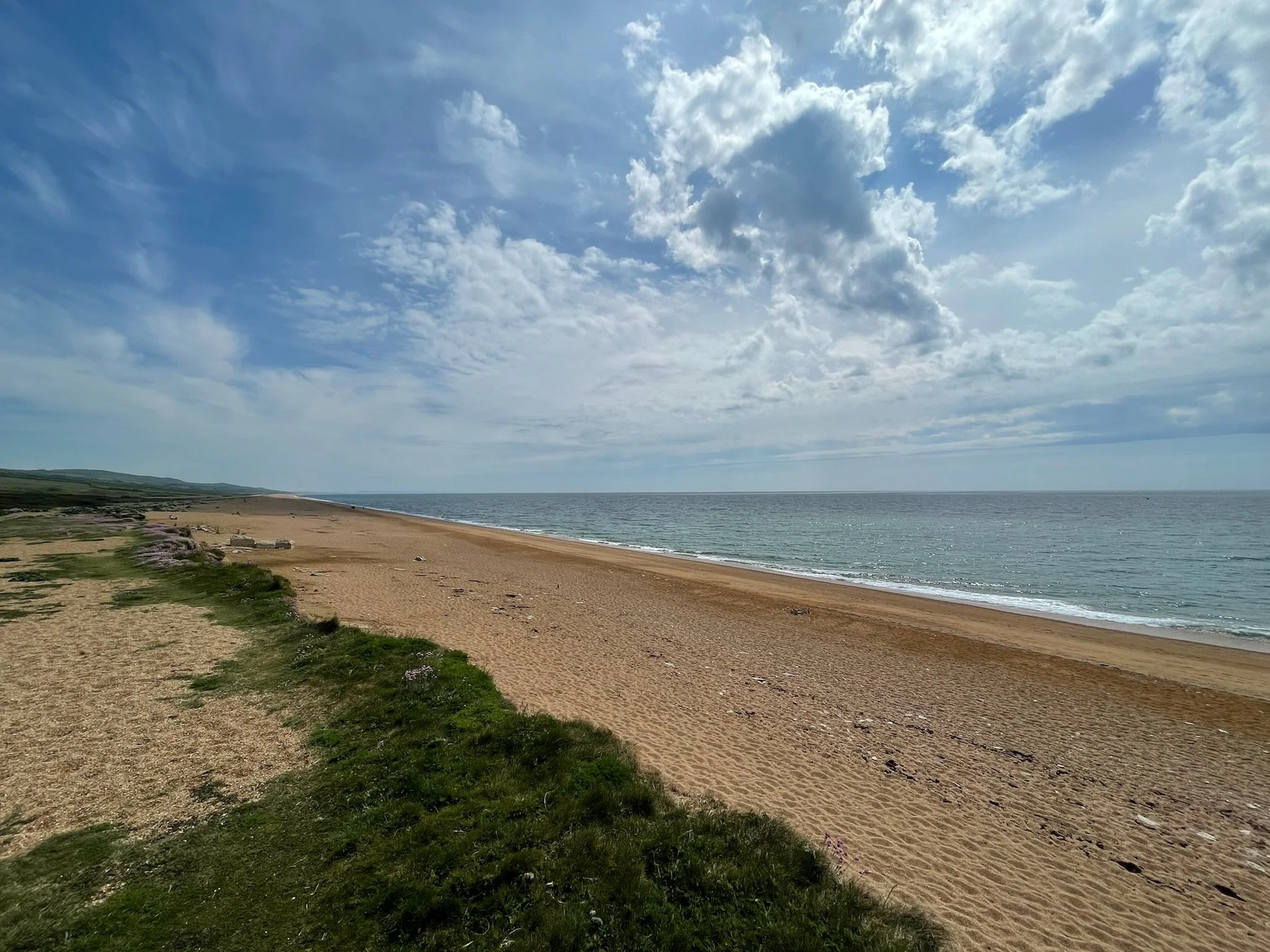 A view of a sandy beach with some grass and rocks along the shoreline, extending toward the ocean with gentle waves. The sky is partly cloudy with blue sky visible.