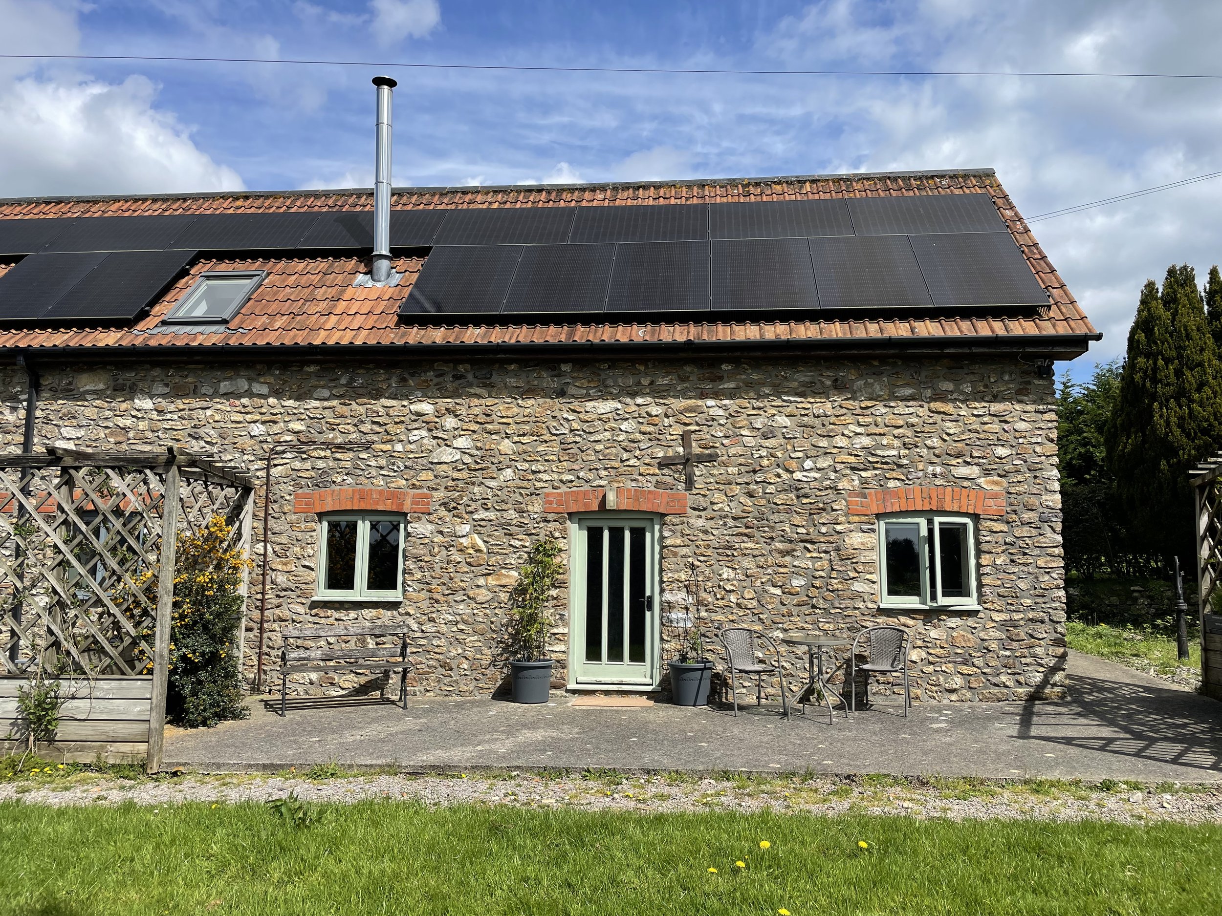 A stone house with a red tile roof fitted with black solar panels. There are windows and a glass door on the ground level, a small garden with plants and outdoor chairs, and a wooden fence in the foreground. The sky is partly cloudy with blue patches.