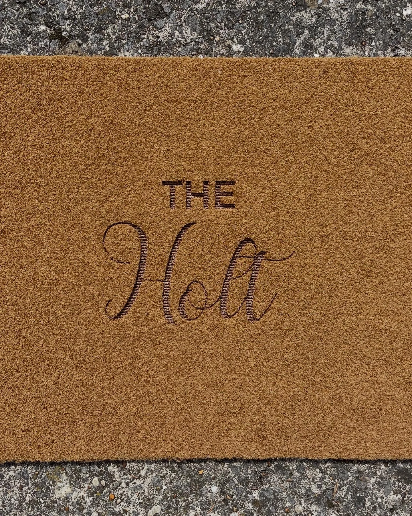 Close-up of a door mat with the words "The Holt" in black on it, lying on a gray and black speckled stone surface.