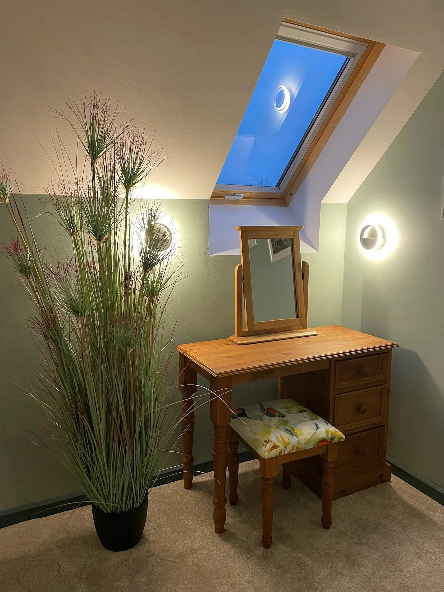 A cozy corner with a wooden vanity table and matching stool with a floral cushion. A tall potted grass plant in black pot is in the foreground. There is a skylight window in the ceiling showing a bright blue sky with a circular LED light fixture near
